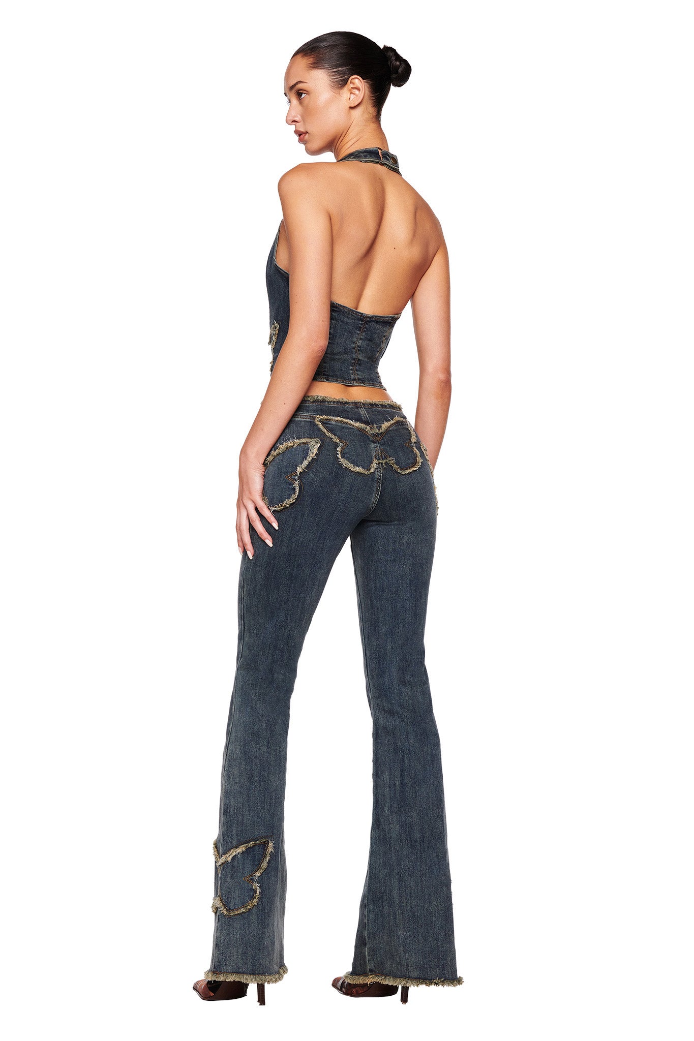 A woman is standing with her back to the camera, wearing a denim outfit. The ensemble features a backless, halter-style top and the SAFFIYAH PANT - BLUE by I.AM.GIA, which are high-waisted stretch denim pants adorned with butterfly cut-outs and frayed edges. She has her hair tied back in a bun.