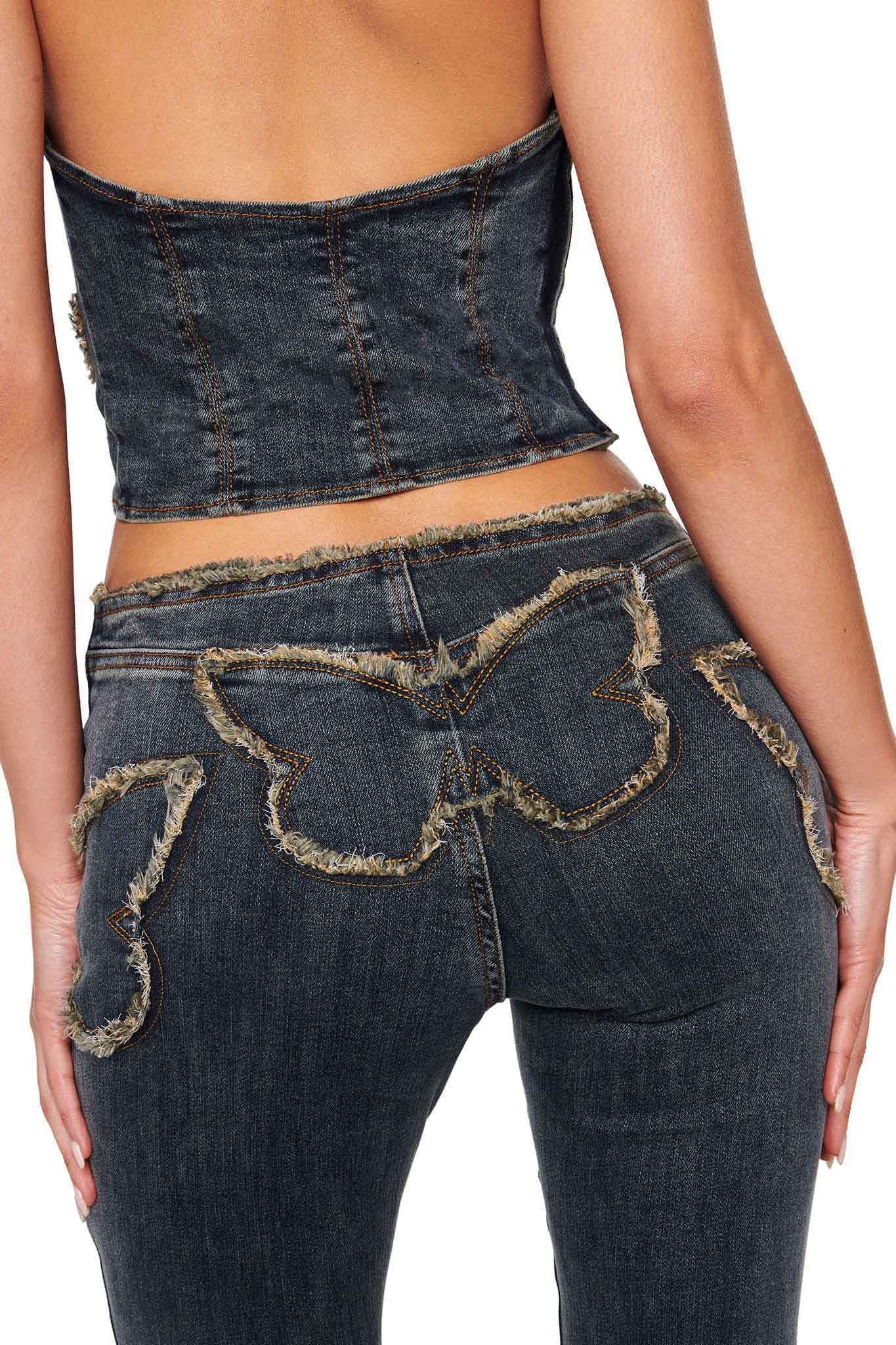 A person wearing a strapless denim top and the I.AM.GIA SAFFIYAH PANT in Denim Blue, flaunting high-waisted, dark wash jeans with flared legs. The back of the jeans features distressed butterfly-shaped patches. Their hands rest on their hips, highlighting the intricate frayed detailing on the stretch denim outfit.