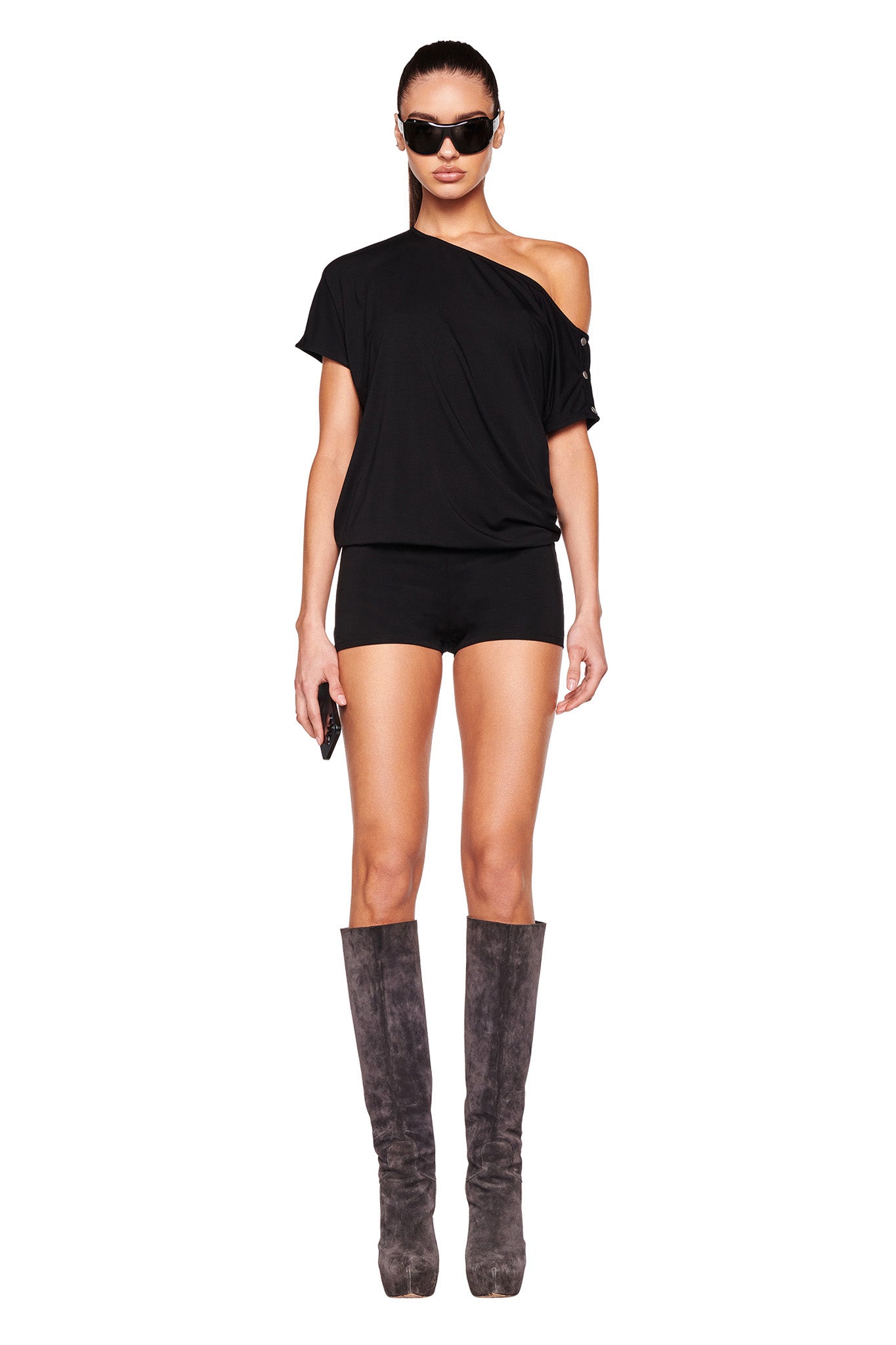 A person stands against a white background wearing an I.AM.GIA EZIA ROMPER - BLACK, knee-high gray boots, and dark sunglasses. They hold a small object in their hand and have a neutral expression.