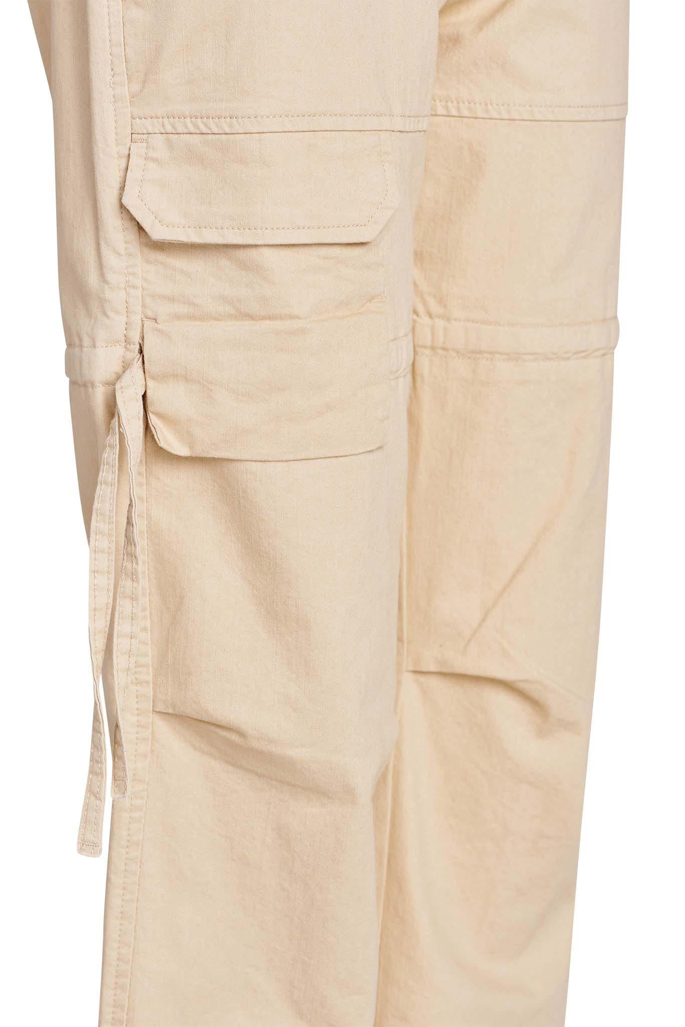 Close-up of the NAIMA CARGO PANT - TAN by I.AM.GIA featuring large side pockets with flaps and adjustable drawstrings. The pants showcase visible stitching and a slightly wrinkled texture, accentuating the rugged design.