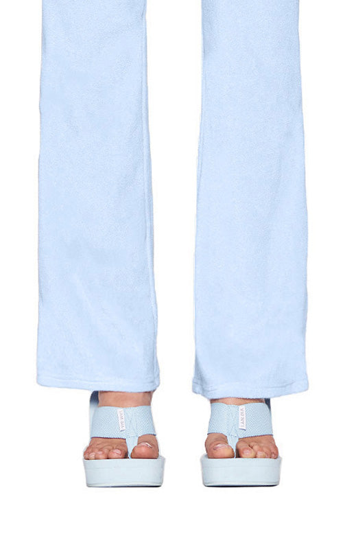A person wearing light blue pants and I.AM.GIA SHELBY FLIP FLOP sandals in baby blue with open toes stands with their feet close together. Only the lower legs and feet are visible in the image.