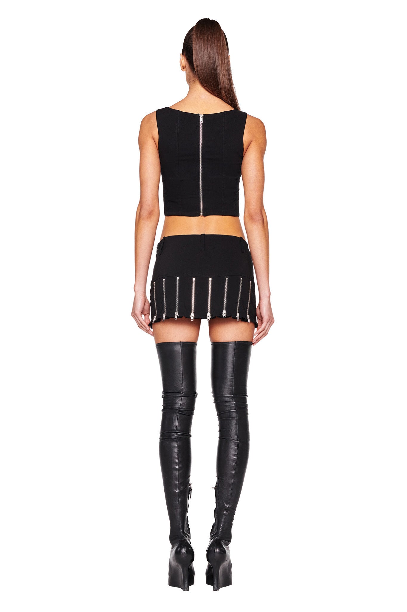 A person with long hair stands facing away from the camera, wearing a black sleeveless crop top and the CLAUDIA SKIRT - BLACK by I.AM.GIA, featuring decorative zippers. They also sport thigh-high, black, high-heeled boots against a plain white background.