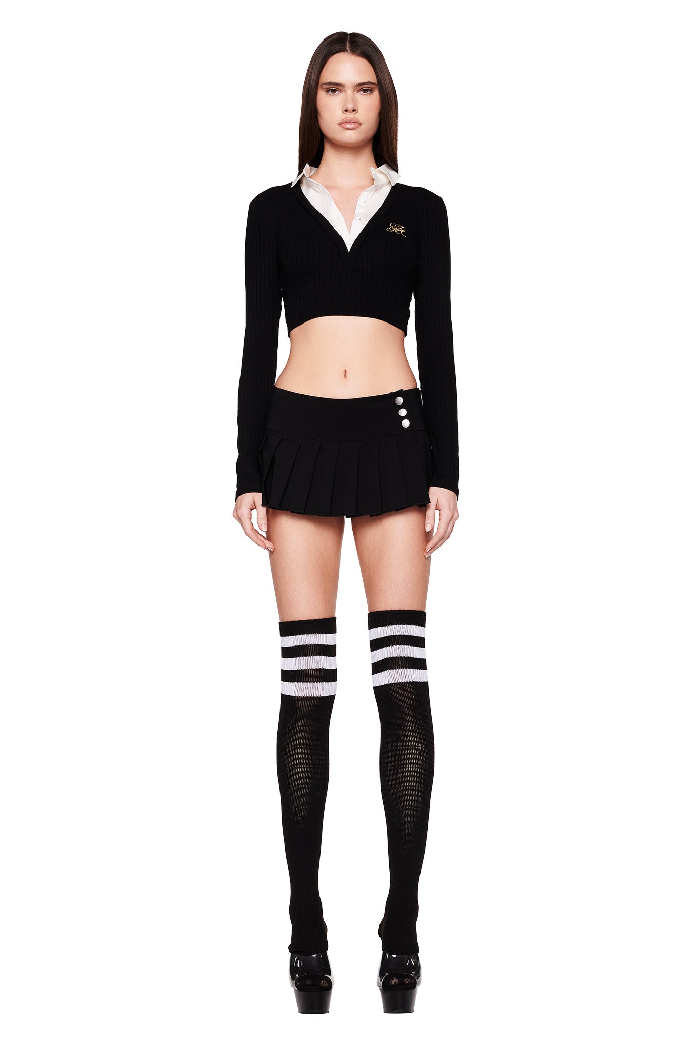 A woman stands wearing the LANDON TOP - BLACK by I.AM.GIA, a cropped sweater with a white collar, paired with a short pleated black skirt featuring buttons on the side, and black thigh-high socks with white stripes. She completes the look with black platform shoes. The background is white.