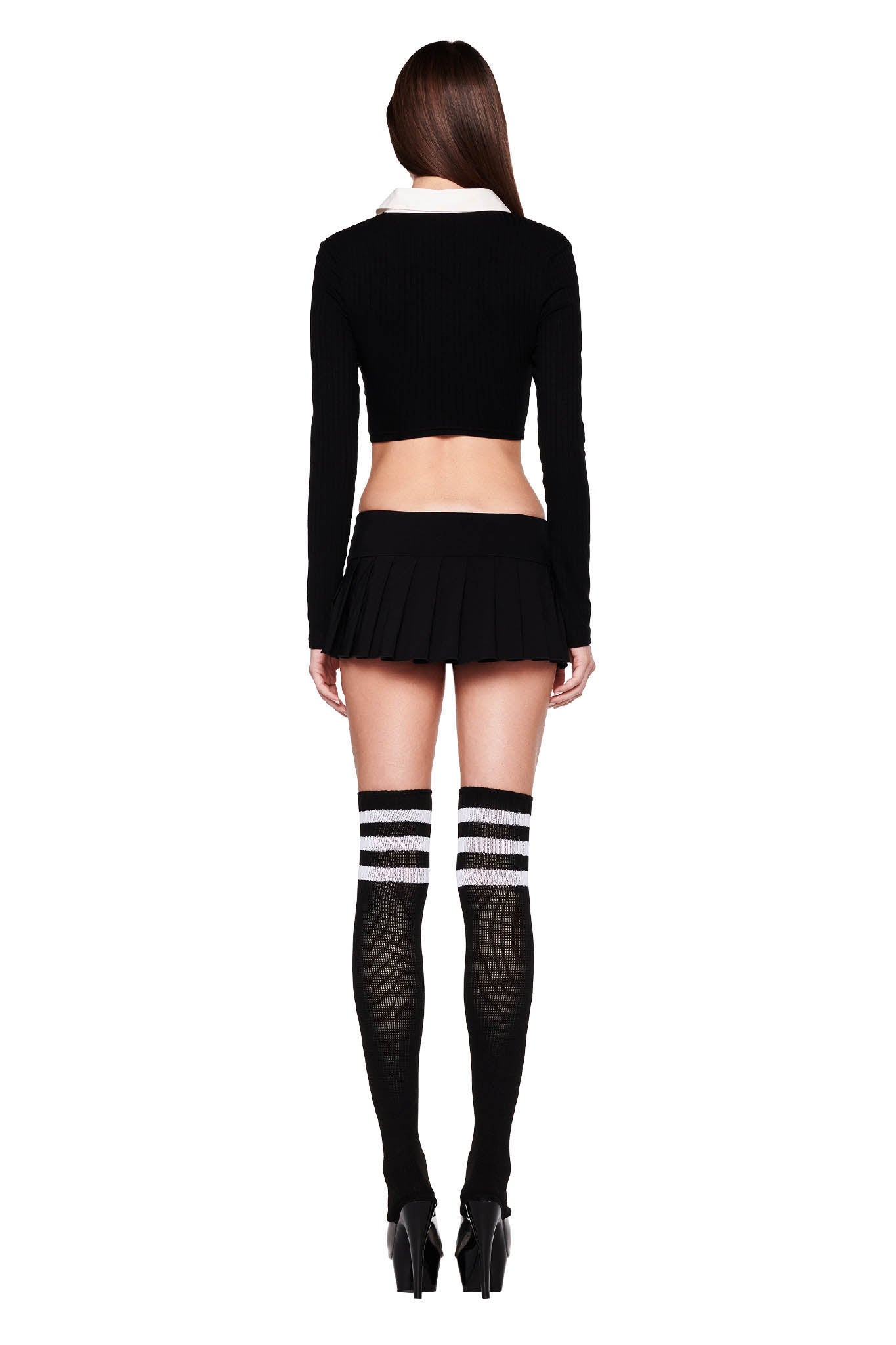 A woman with long dark hair faces away from the camera, showcasing her I.AM.GIA LANDON TOP in black, a short pleated skirt, and black thigh-high stockings with white stripes. She stands confidently in black high-heeled shoes against a plain white background.