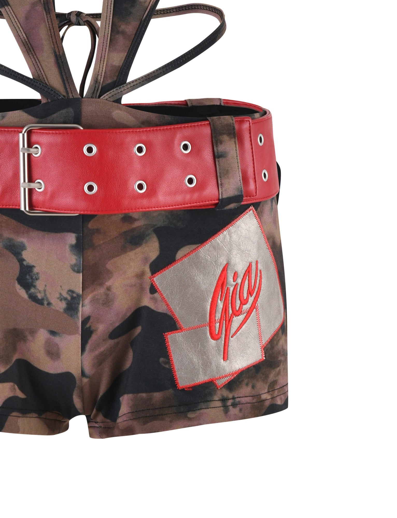 A close-up of the LIANNA ROMPER - GREEN : CAMO by I.AM.GIA, featuring camouflage shorts with a large red belt adorned with silver eyelets. The shorts boast a red and gray patch with the name "Gia" stitched in red on the right side. Star patches accentuate the design, and background belts suggest a harness or strappy look.