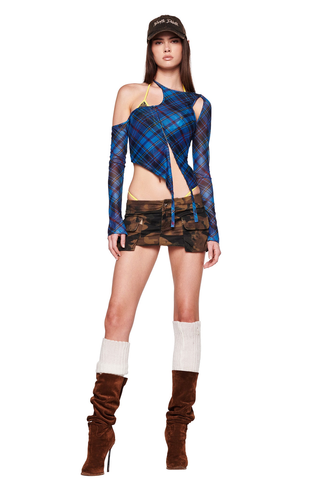 A woman stands wearing a unique I.AM.GIA outfit, featuring the ANAFER TOP - BLUE : PLAID with asymmetrical cut-out detailing, camouflage shorts, brown knee-high boots paired with white leg warmers, and a brown baseball cap. She poses confidently, highlighting the eclectic fashion ensemble.