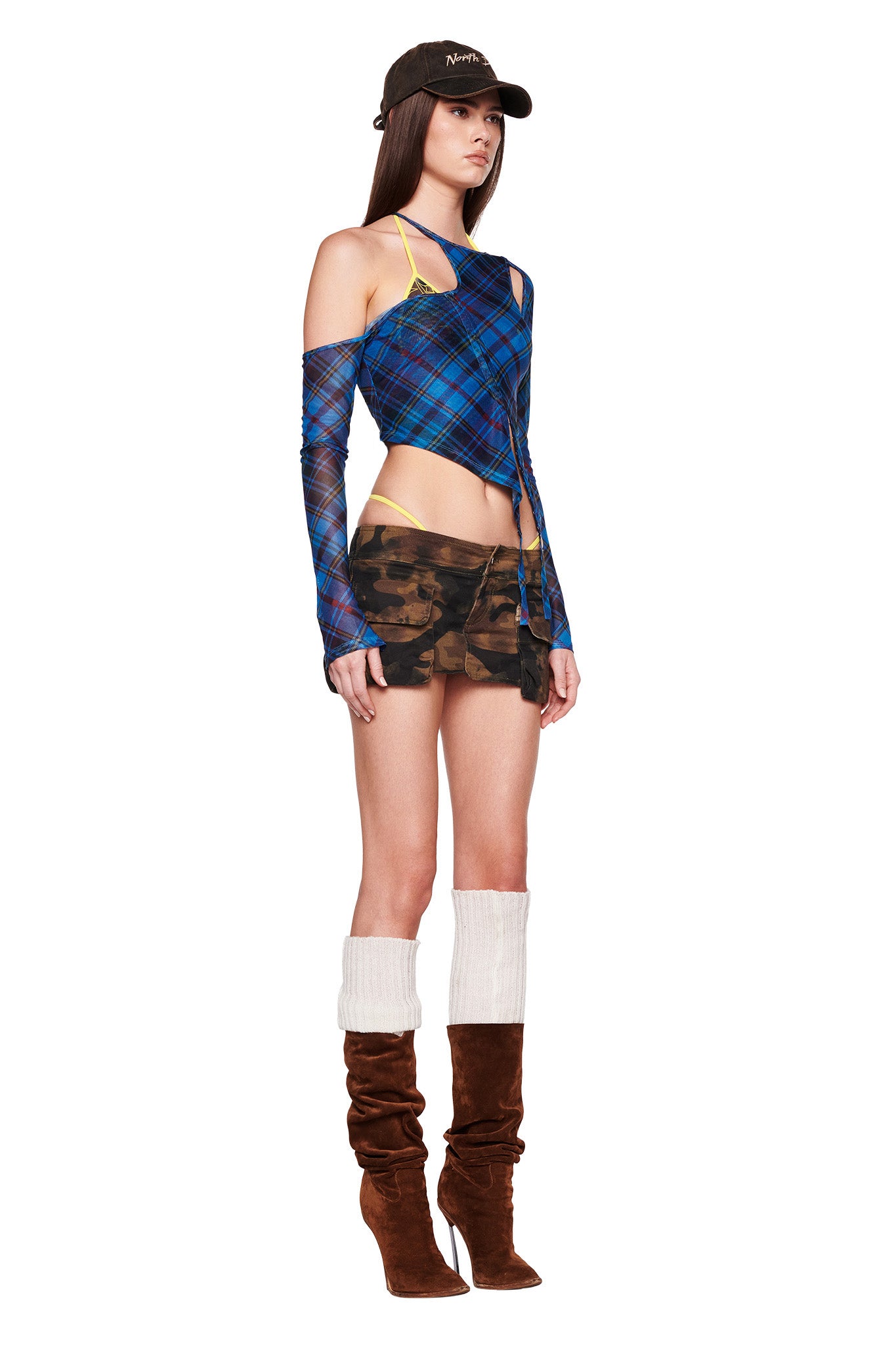 A woman stands in a side view, showcasing her ensemble with a black cap, the ANAFER TOP - BLUE : PLAID by I.AM.GIA, a camo mini skirt, and brown knee-high boots with white leg warmers. The background is plain white.