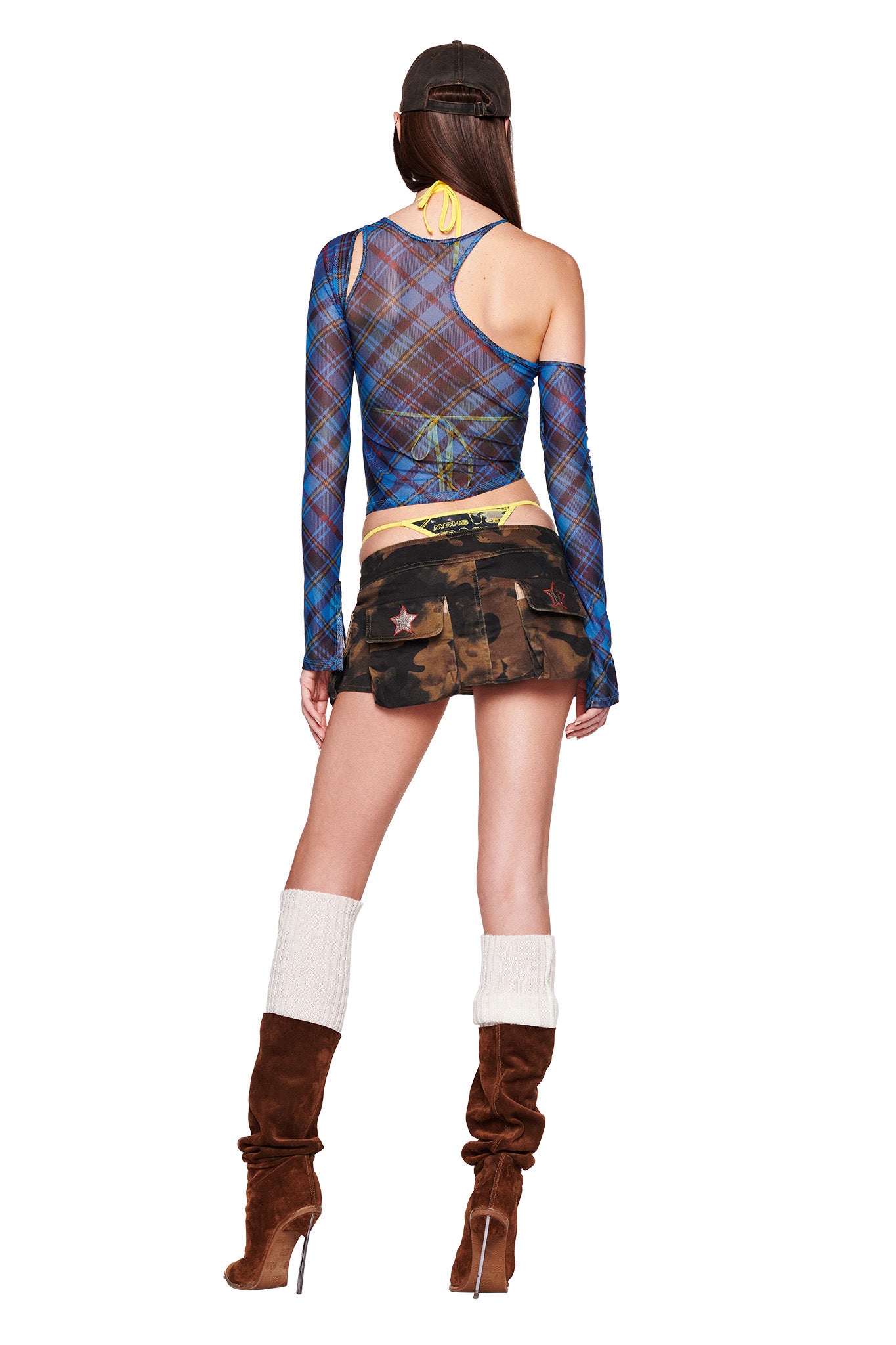 A model is standing with her back to the camera, wearing the ANAFER TOP - BLUE : PLAID from I.AM.GIA, which features an asymmetrical design with one sleeve. She pairs it with a camouflage miniskirt also from I.AM.GIA, brown high-heeled boots accompanied by white leg warmers, and a black baseball cap. Her long hair cascades down her back.