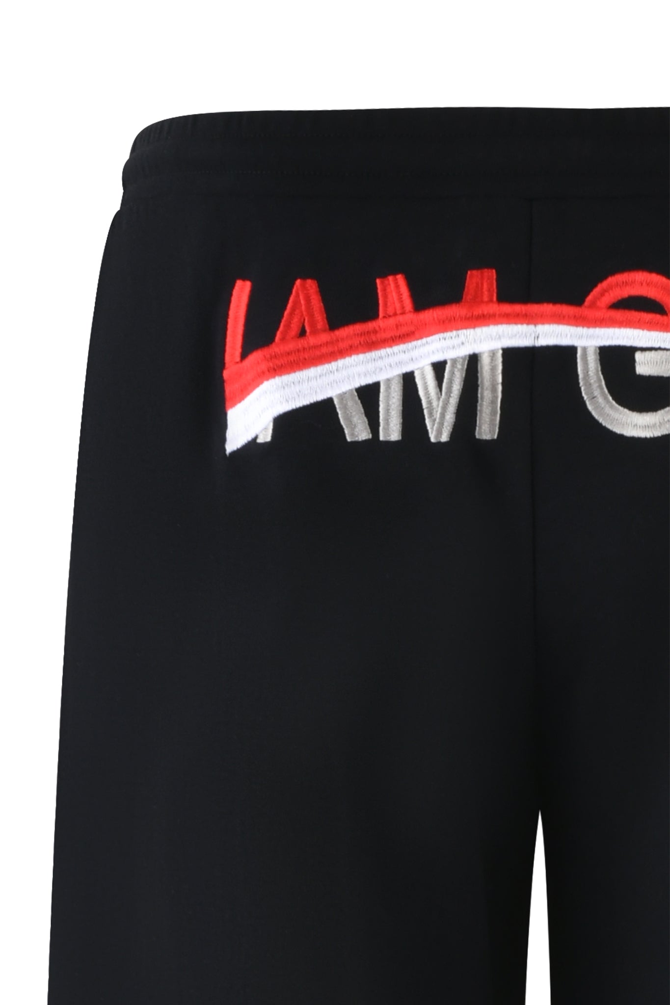 Close-up of the JUSTINE PANT - BLACK by I.AM.GIA, showcasing baggy track pants with an elastic waistband. The partially visible GIA embroidery transitions from red "IAM" to silver "ORG" in red, white, and silver thread on a sleek black background.