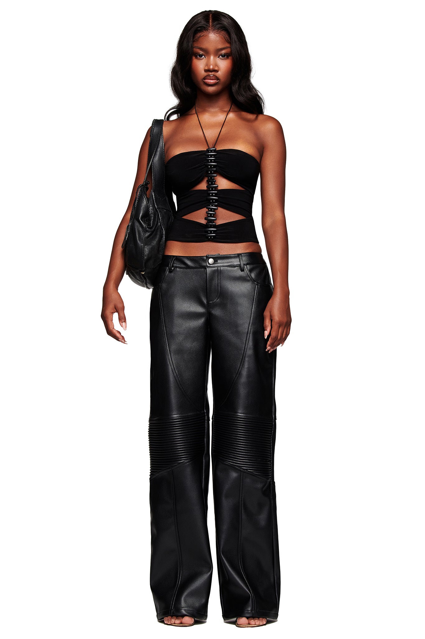 A woman with long, wavy hair and a neutral expression faces forward against a white background, wearing the I.AM.GIA KRISTIE PANT in black styled with a black strapless cut-out top and a black shoulder bag.