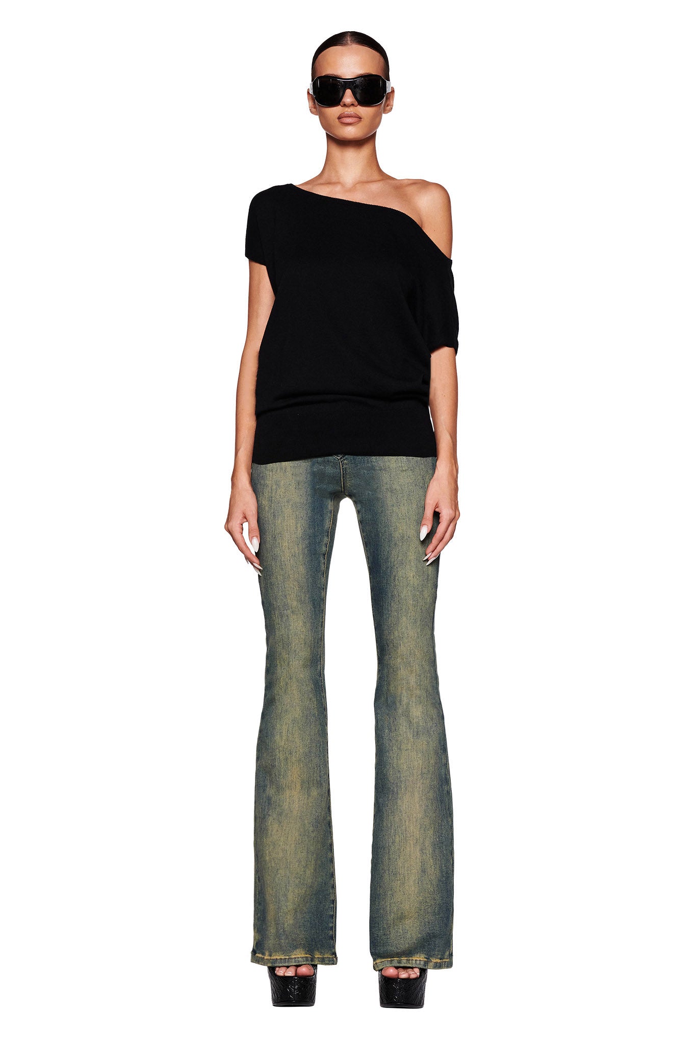 A person stands against a white background wearing the GISELLE SHORT SLEEVE TOP - BLACK by I.AM.GIA, blue flared jeans, black platform shoes, and large black sunglasses. Their hair is pulled back, and they have a confident posture with arms relaxed by their sides.