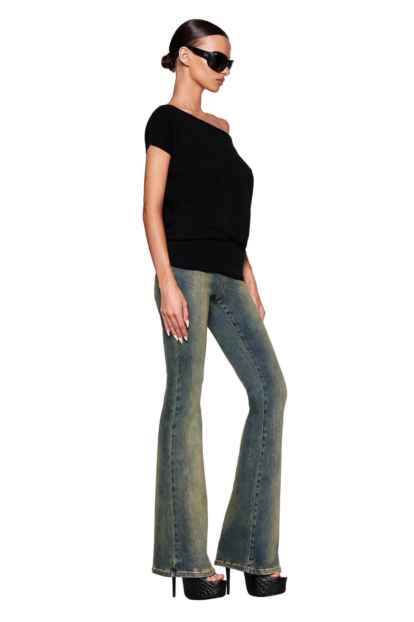 A woman stands against a white background, wearing sunglasses and the GISELLE SHORT SLEEVE TOP - BLACK by I.AM.GIA, made from fully fashioned knit fabrication. She pairs it with faded blue flared jeans and black platform heels. Her hair is styled into a low, sleek bun.