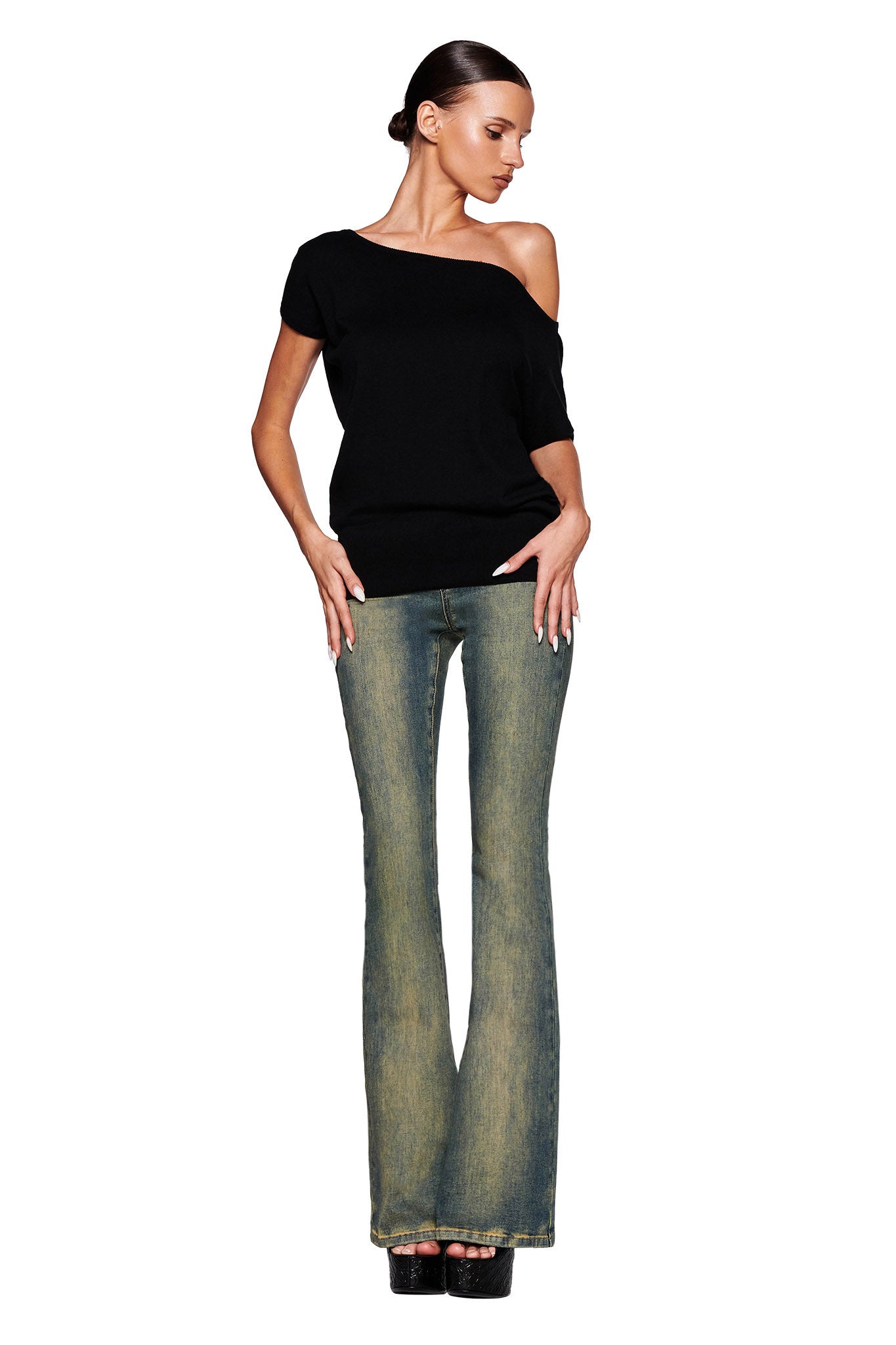 A woman with dark hair tied back is posing against a white background. She is wearing an I.AM.GIA GISELLE Short Sleeve Top in black, faded bell-bottom jeans, and black platform shoes. Her hands are resting on her hips, and she is looking down to the left.