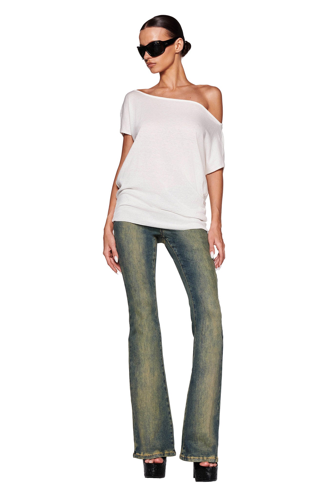 A woman wearing dark sunglasses, the GISELLE SHORT SLEEVE TOP in white by I.AM.GIA with ribbed detail, and faded blue flare jeans stands confidently with one hand in her pocket and the other relaxed by her side. Her hair is neatly tied back, and she is wearing black platform shoes. The background is plain white.