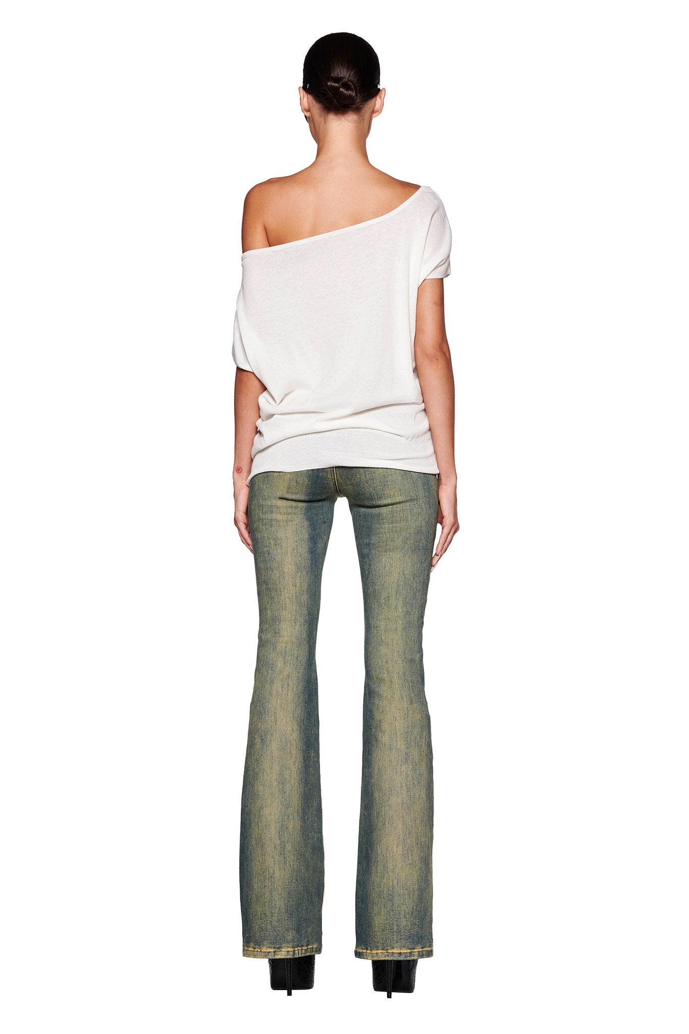 A person with dark hair, tied back, is standing with their back facing the camera. They are wearing the I.AM.GIA GISELLE SHORT SLEEVE TOP in white with ribbed detail, fitted blue flare jeans, and black platform shoes. The background is plain white, highlighting the outfit.