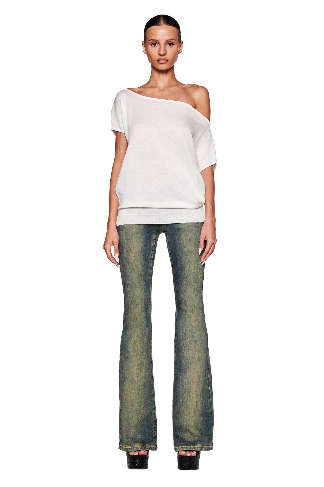 A woman stands against a white background wearing the GISELLE SHORT SLEEVE TOP - WHITE by I.AM.GIA, featuring off-the-shoulder design and ribbed detailing. She pairs it with faded flare jeans and black platform shoes. Her hair is pulled back and she faces the camera with a neutral expression.