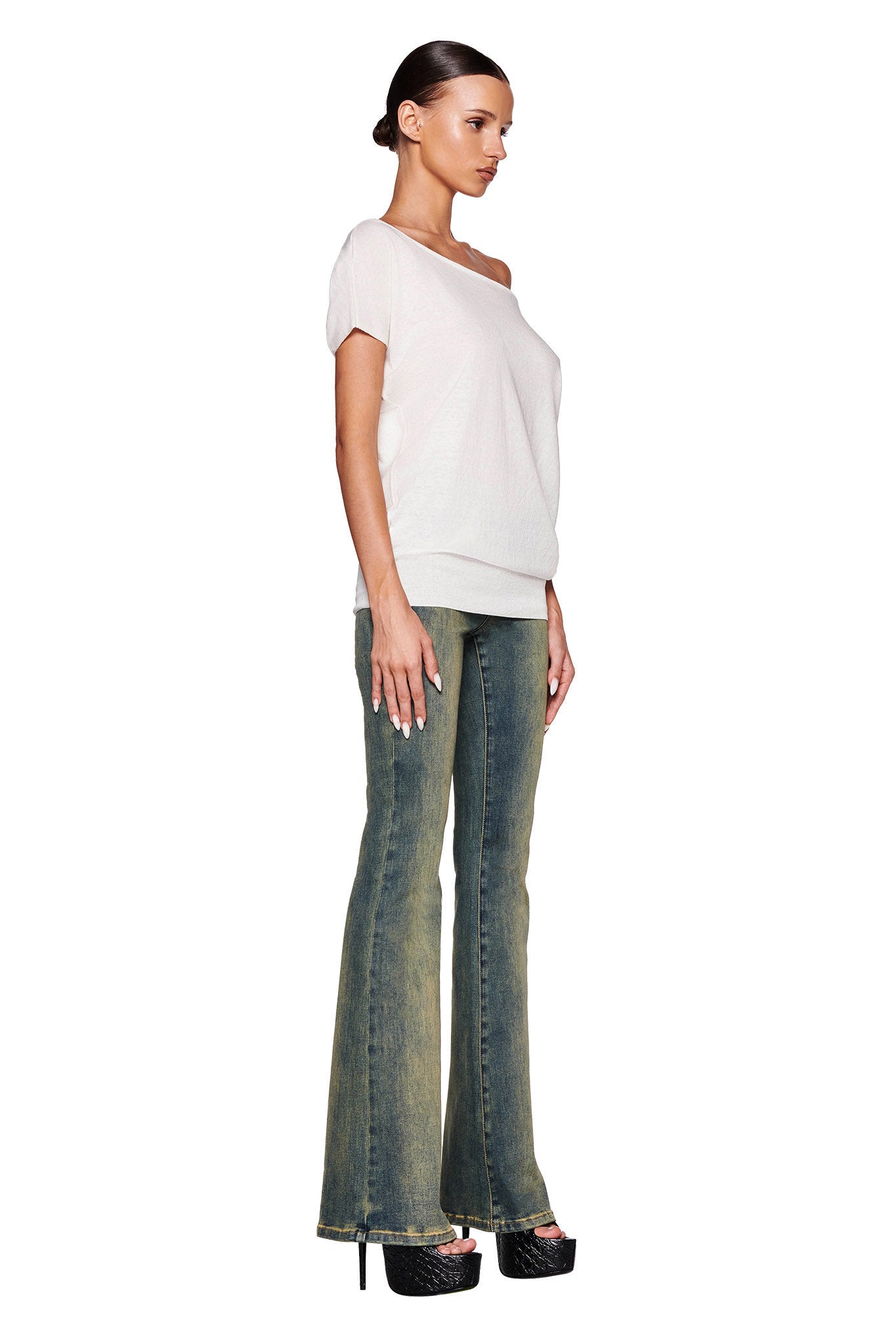 A woman stands against a white background, facing sideways. She wears the GISELLE SHORT SLEEVE TOP in white from I.AM.GIA, paired with blue high-waisted flare jeans and black platform heels. Her hair is styled in a low, sleek bun.