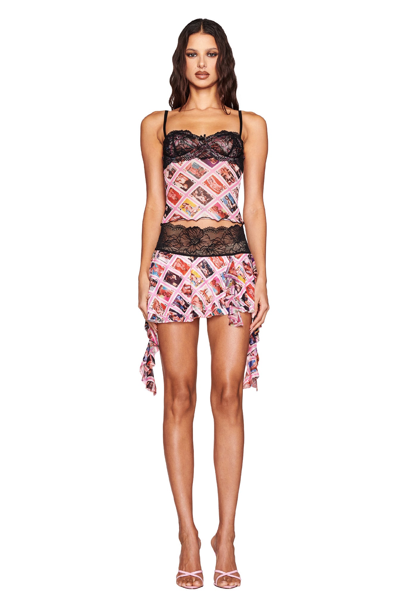 A woman wears the I.AM.GIA SILO MINI SKIRT - MULTI: GIA FACE CARD, featuring a postage stamp pattern with black lace and an asymmetrical mini design. Paired with a sleeveless top and strappy high heels, she stands against a white background.