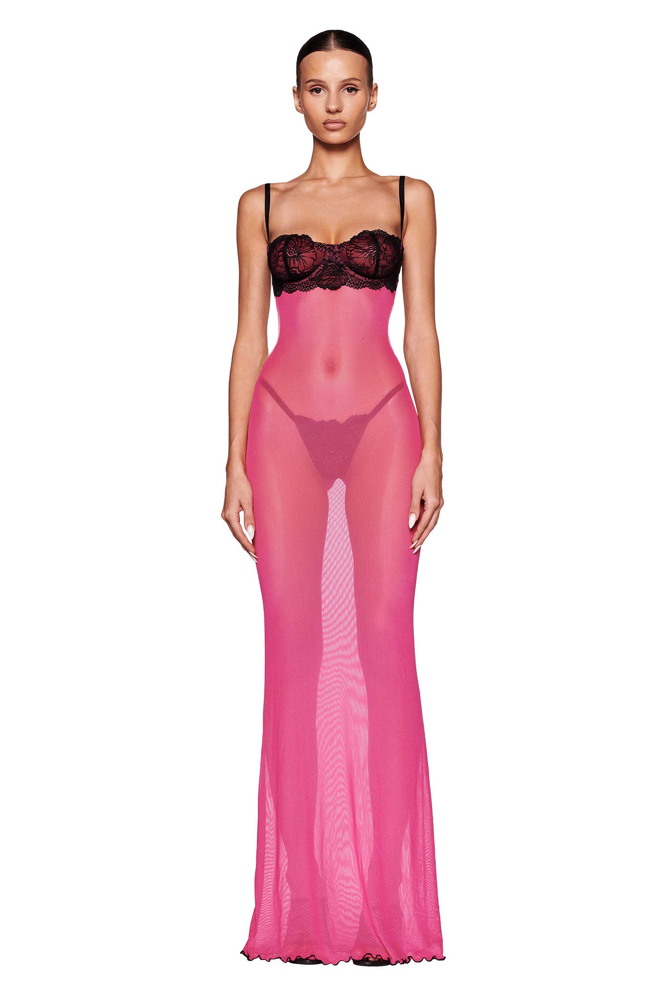 A person stands wearing the SILO MAXI DRESS - PINK by I.AM.GIA. The hot pink dress features sheer mesh material with black lace detailing on the bodice. It is a strapless maxi-length dress enhanced by thin straps over the shoulders. The individual exudes poise and confidence, with their hair neatly pulled back.