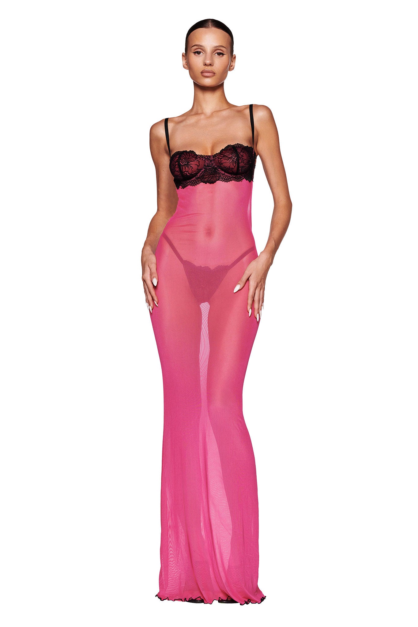 A person stands confidently modeling the SILO MAXI DRESS - PINK : HOT PINK by I.AM.GIA. The sheer mesh dress, accentuated with black lace details, flows elegantly to the floor in a maxi length style. Featuring spaghetti straps and a fitted bodice, the model rests their hands gently on their thighs against a pristine white background.