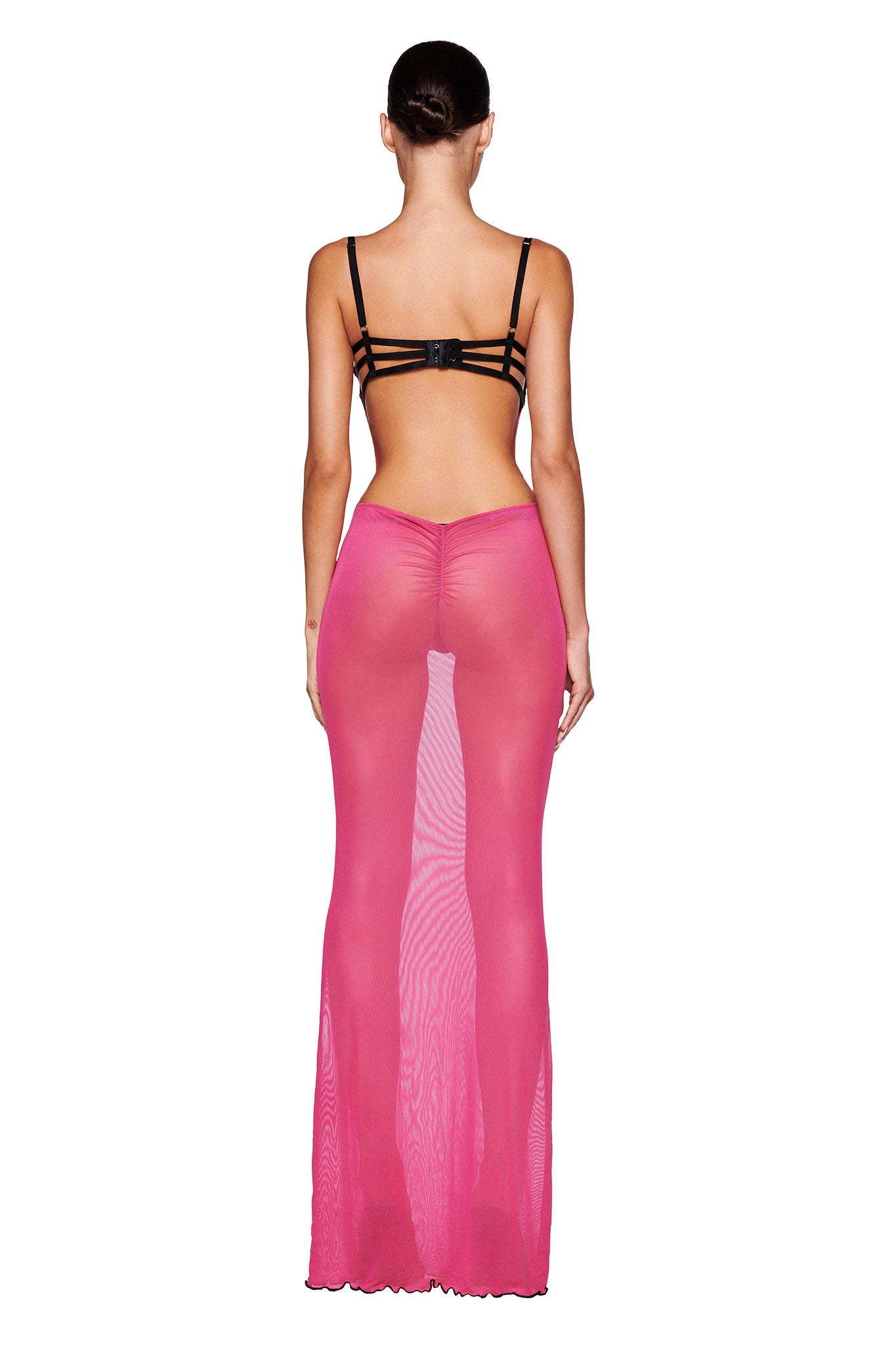 A person with short dark hair is standing with their back to the camera, wearing a black strappy bra and the SILO MAXI DRESS in HOT PINK by I.AM.GIA. The sheer, maxi-length dress reveals the form and silhouette of the body underneath.