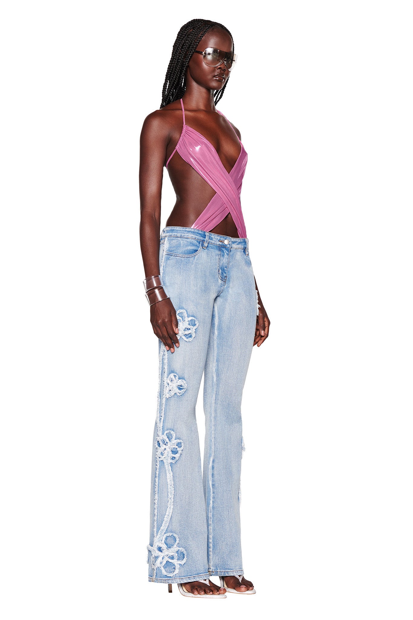 A woman with long braided hair stands in a confident pose wearing a stylish, deep pink I.AM.GIA halter top and the TYLA JEAN - BLUE by I.AM.GIA, light blue denim bell-bottom jeans with floral appliqués. She accessorizes with metallic bracelets, sunglasses, and high-heeled sandals.