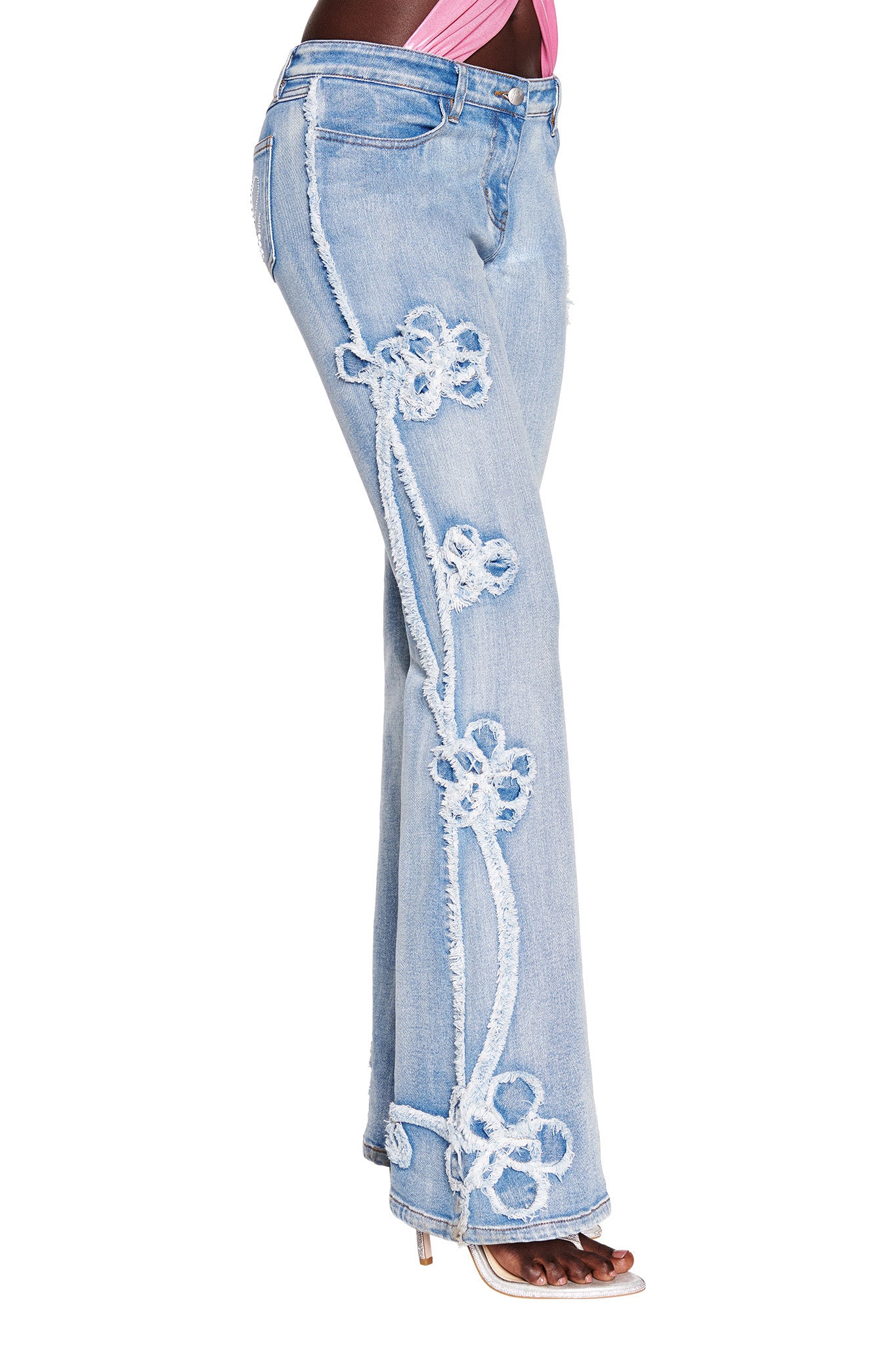 Side view of a person wearing the TYLA JEAN - BLUE : DENIM BLUE from I.AM.GIA, decorated with frayed floral appliqués along the outer seam. The person stands with legs slightly apart, showcasing the jeans paired with a pink top and high-heeled sandals.