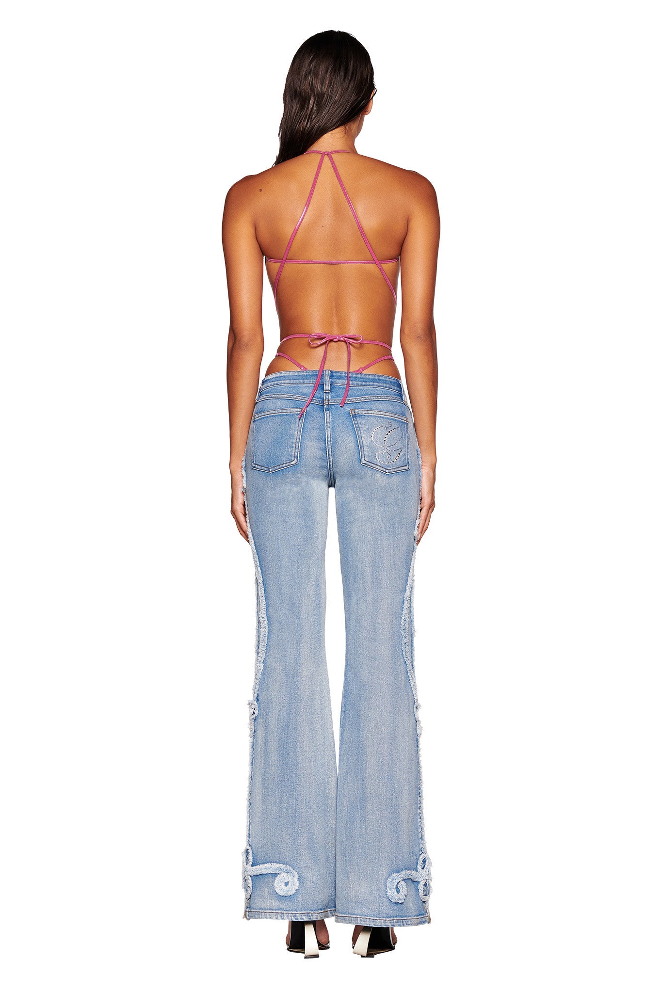 A woman is standing with her back facing the camera, dressed in stylish I.AM.GIA TYLA JEAN - BLUE featuring decorative stitching on the hems and pockets, a backless top with thin criss-cross straps, and black high heels. Her long hair flows down her back.
