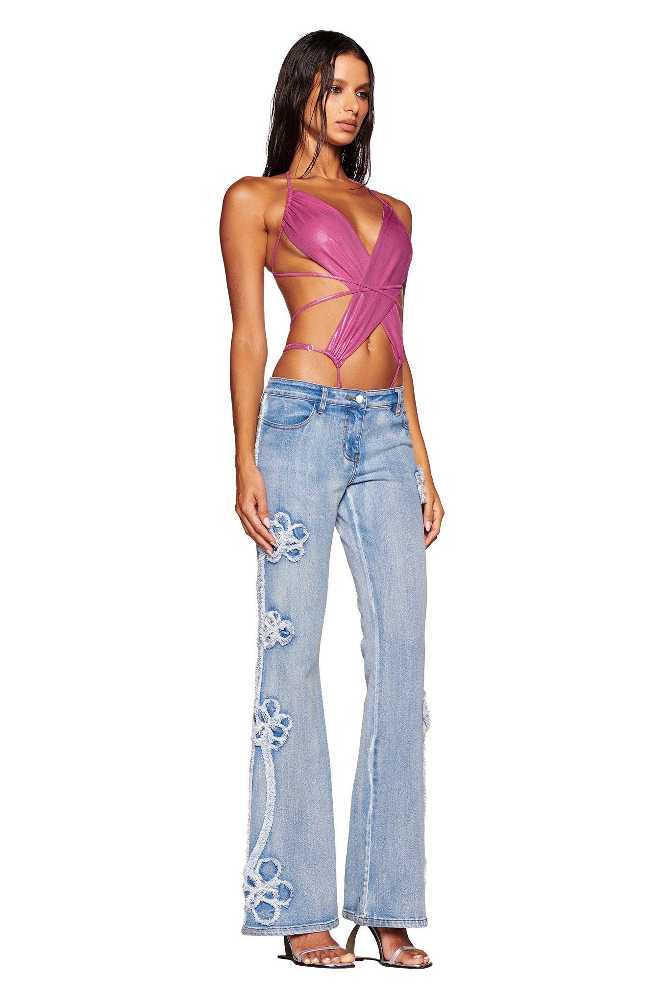 A woman poses wearing a strappy, fuchsia I.AM.GIA top with a plunging neckline and crisscross design. She pairs it with the TYLA JEAN - BLUE by I.AM.GIA, featuring light blue high-waisted denim adorned with large white flower patterns down the sides. She has long straight hair and is wearing open-toe heels.