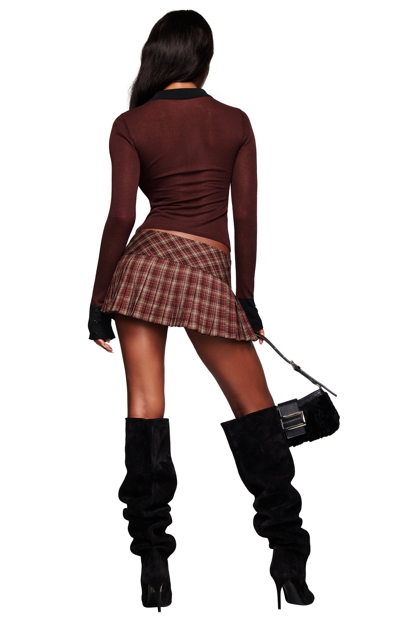 A person stands facing away, wearing the I.AM.GIA DAVINA TOP in brown, a short pleated plaid skirt, tall black heeled boots, and holding a small black handbag.