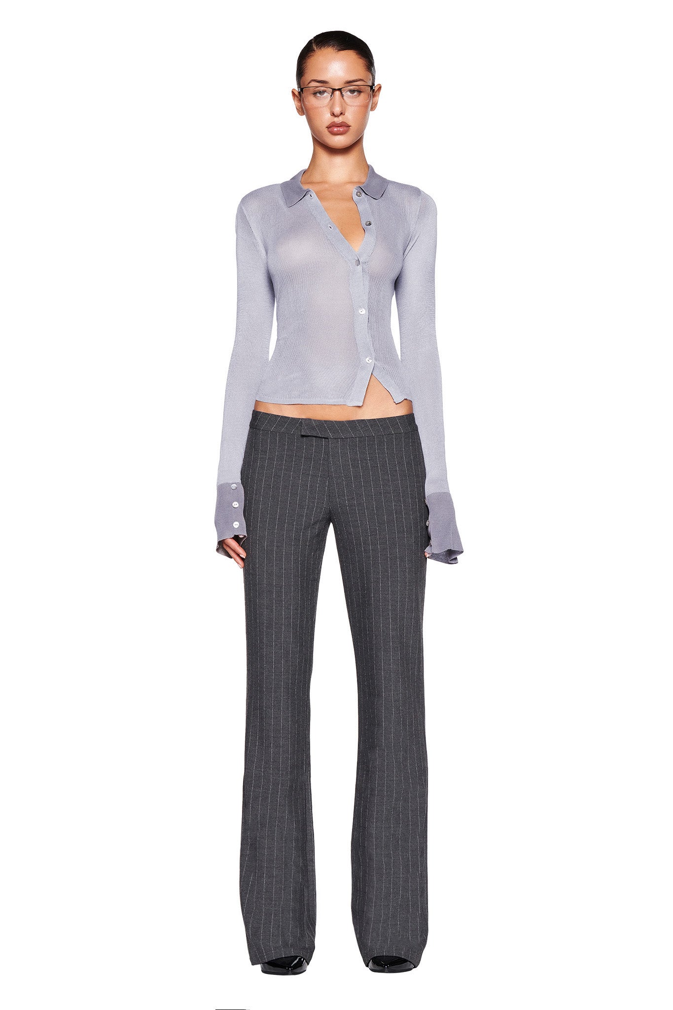 A person stands against a white background, wearing the I.AM.GIA DAVINA TOP in grey with the bottom button undone and gray pinstripe trousers. They also wear glasses, and their hair is pulled back. The grey long sleeve top complements their neutral expression perfectly.