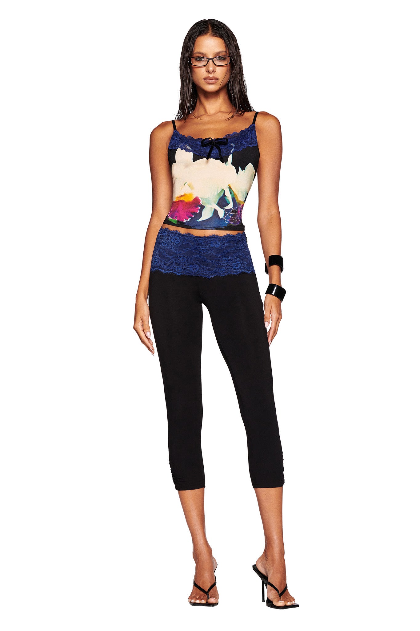A woman with long dark hair, wearing glasses, is dressed in a custom graphic ADELLA TOP - BLACK : FLORAL : BLACK ORCHID by I.AM.GIA with blue lace, black capri leggings, and black high-heeled sandals. She stands against a plain white background.
