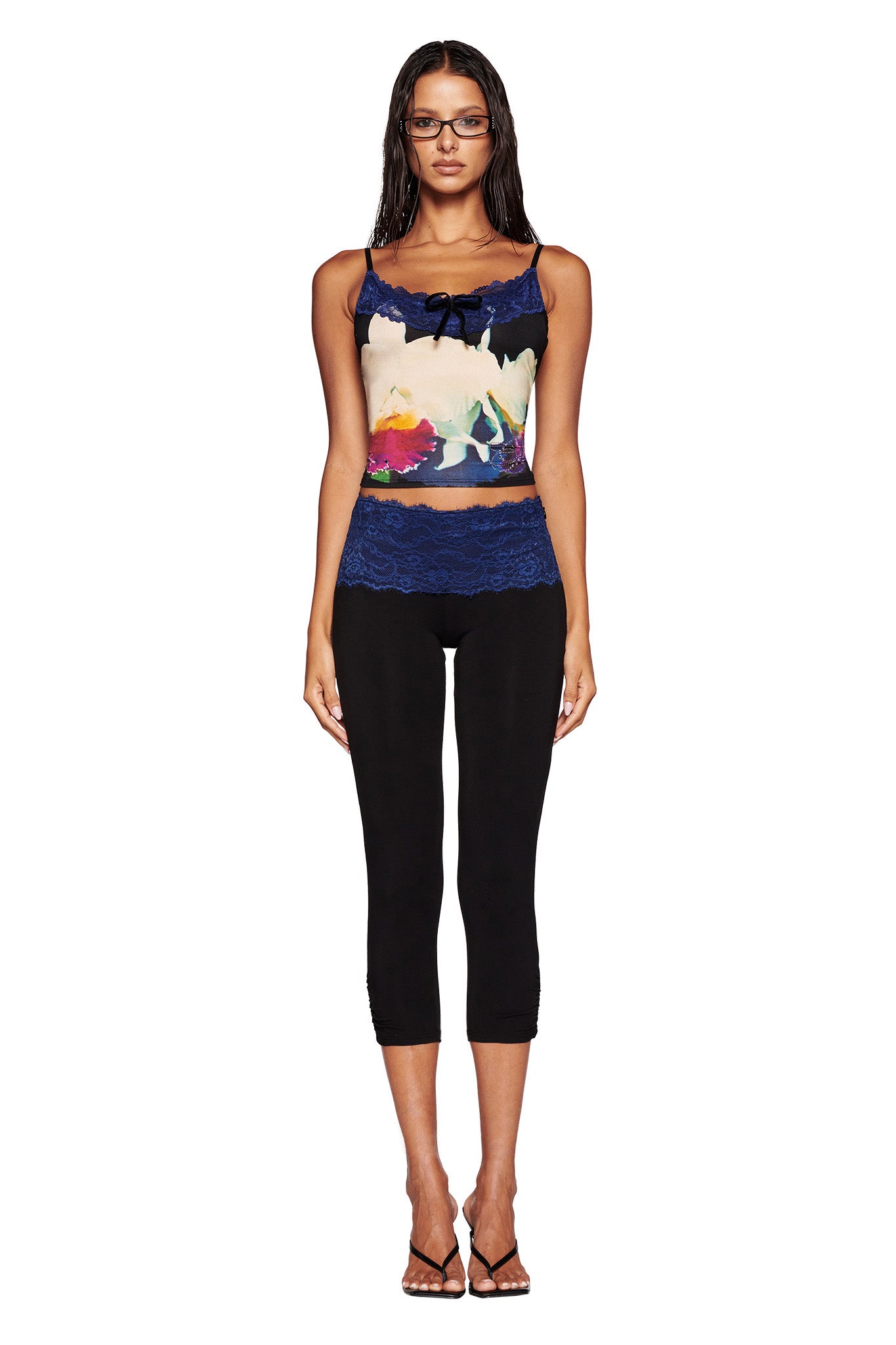 A woman stands facing the camera, wearing a colorful floral tank top with lace details and black capri pants from the I.AM.GIA collection. She has long, straight hair and wears glasses, standing in a neutral pose with her arms at her sides. She completes her look with black heeled sandals.