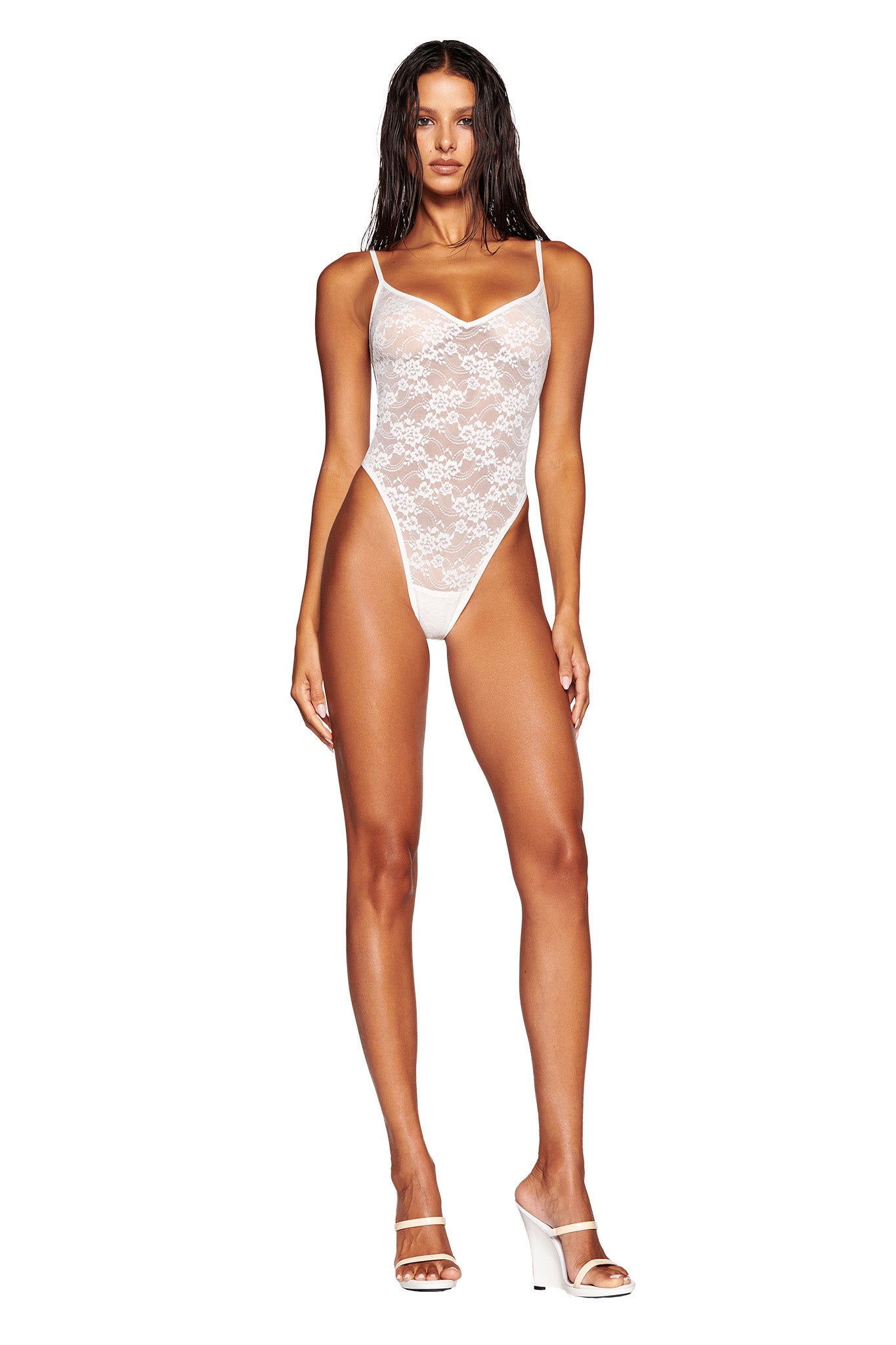 A person stands against a plain white background, wearing the PERLA BODYSUIT - WHITE by I.AM.GIA and white heeled sandals. They have long, wet hair and a neutral expression.