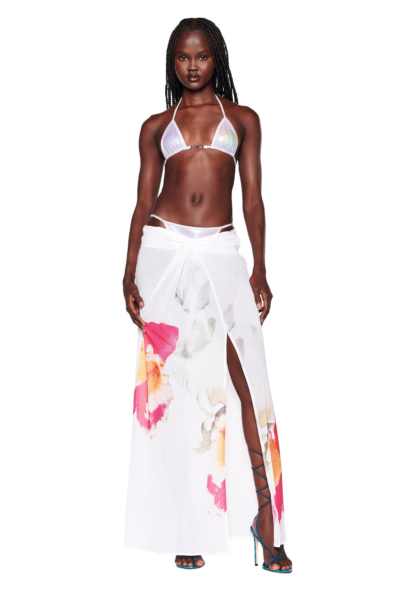 A woman is modeling an I.AM.GIA two-piece outfit, featuring a shiny silver bikini top paired with the PURA MAXI SKIRT - WHITE : FLORAL. She faces forward with braided hair, wearing teal high-heeled sandals, and stands against a plain white background.
