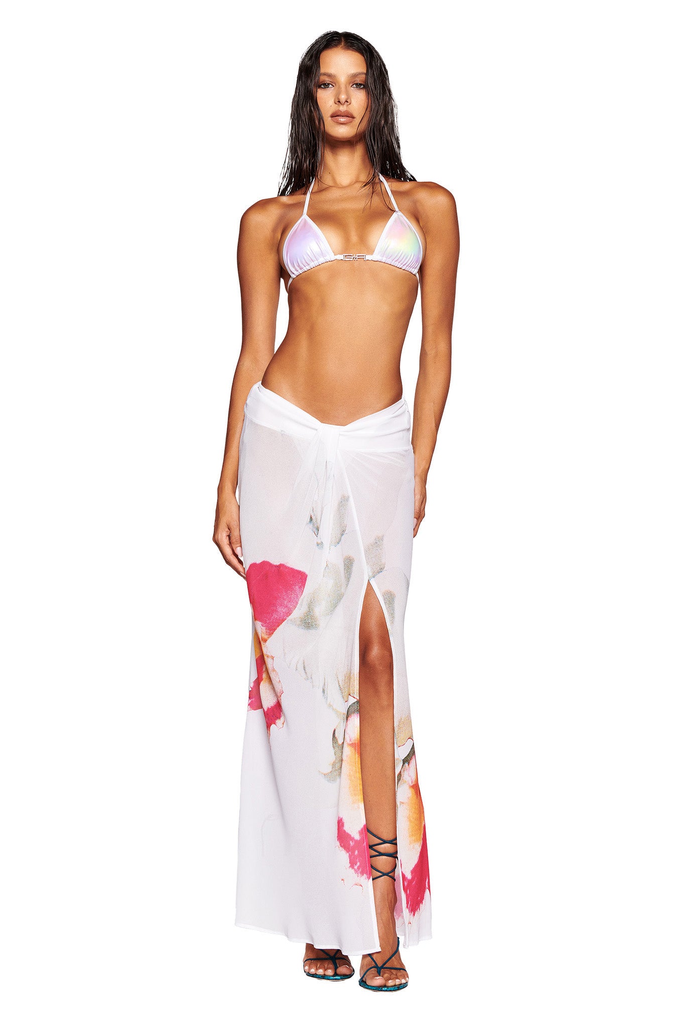 A woman stands wearing an I.AM.GIA colorful string bikini top and the PURA MAXI SKIRT - WHITE : FLORAL, a sheer white wrap skirt adorned with floral designs. She has long, wavy hair and is posing confidently, looking straight ahead. Strappy sandals complete her look.