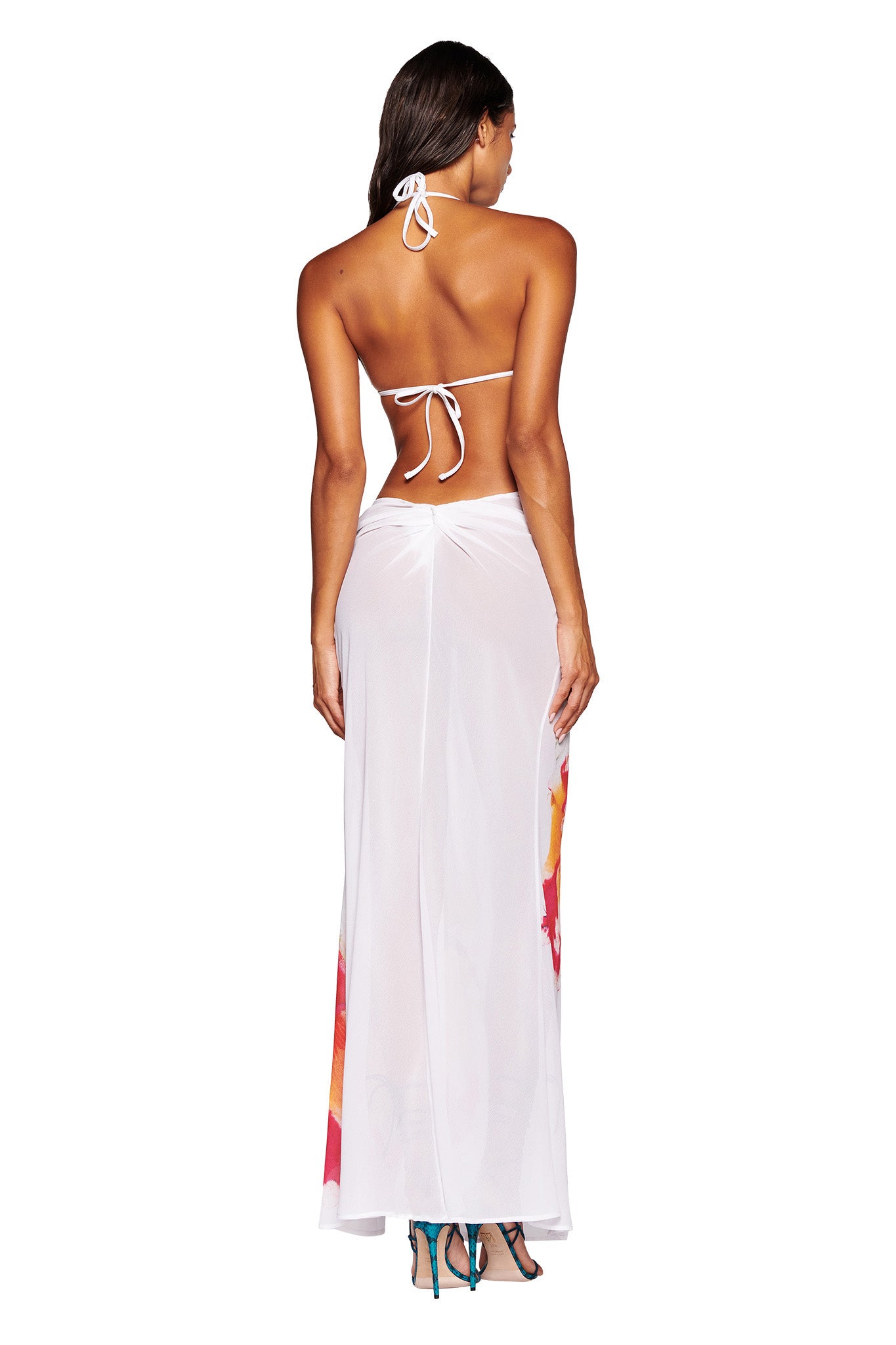 A woman is seen from behind wearing a white halter neck bikini top and the I.AM.GIA PURA MAXI SKIRT - WHITE : FLORAL, which features sheer fabric with floral patterns on the sides. She has long hair and stands with her weight shifted to one hip, showcasing her blue high-heeled shoes. Her chic ensemble exudes the edgy sophistication of I.AM.GIA.