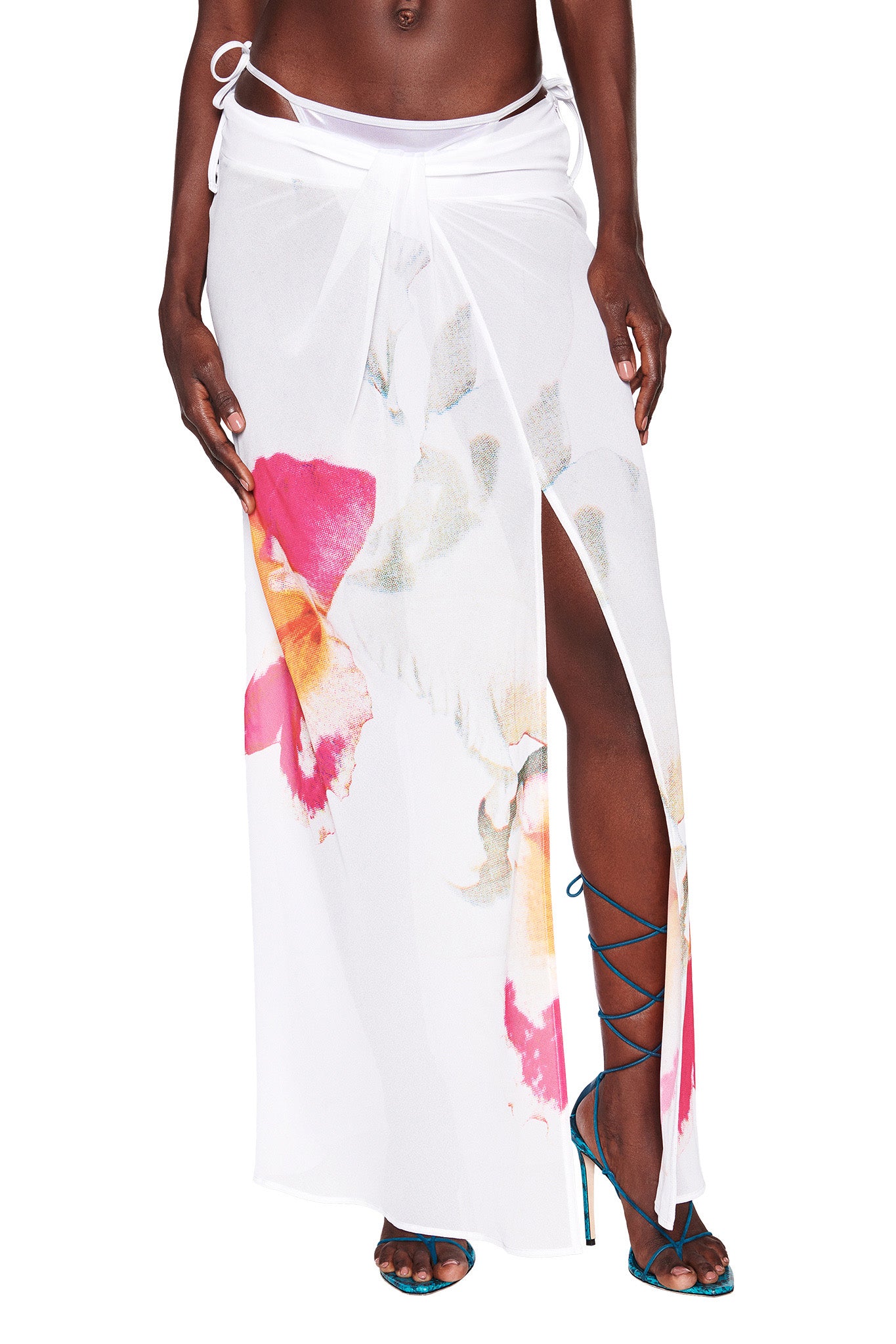 A person is wearing the PURA MAXI SKIRT - WHITE: FLORAL from I.AM.GIA, a stunning sheer wrap skirt adorned with bold floral prints in shades of pink, yellow, and green. The skirt features a high slit that reveals one leg. Paired with strappy blue high-heeled sandals, the ensemble exudes elegance and style.