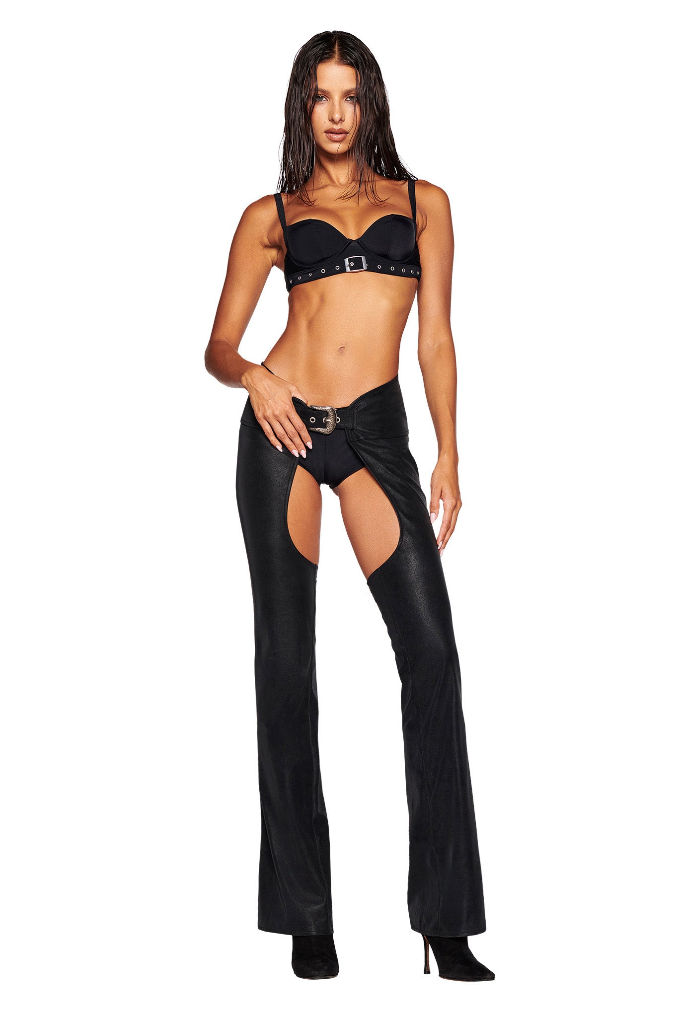 A woman with wet, wavy hair stands confidently. She is dressed in a black bra with buckles and the ARIZONA CHAP PANT in black by I.AM.GIA, paired with black pointed-toe heels and flaunting a flared silhouette. Her left hand rests on her thigh while her right arm hangs by her side. The background is white.