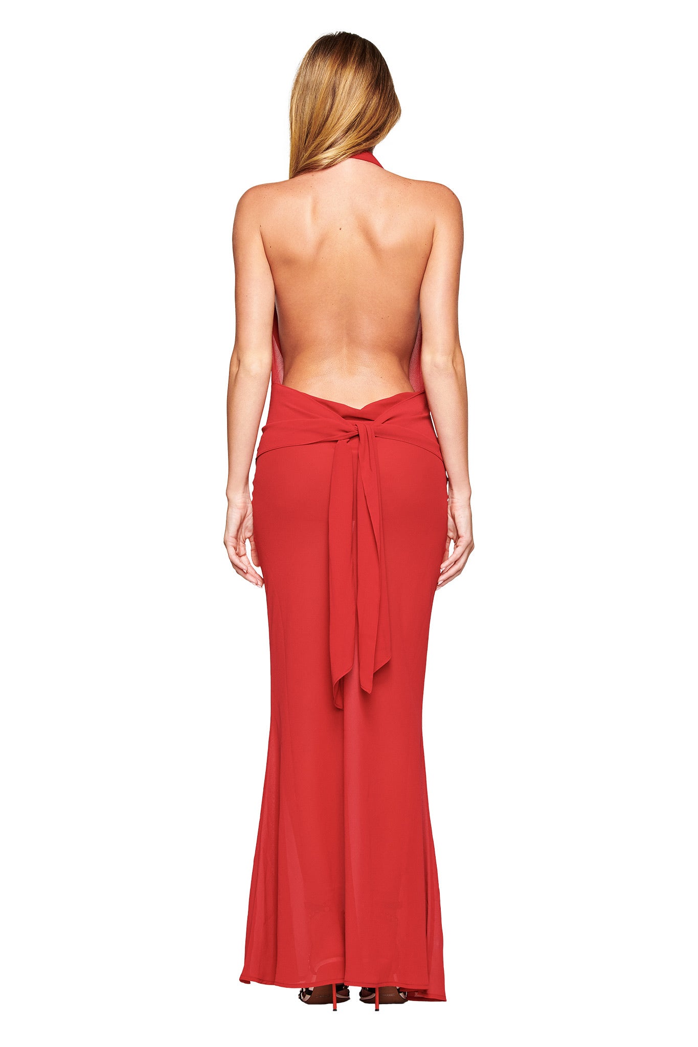 A woman with long blonde hair stands with her back to the camera in the I.AM.GIA KAHLO MAXI DRESS - RED, a backless georgette gown featuring a wide sash tied in a bow at the waist, set against a white background.