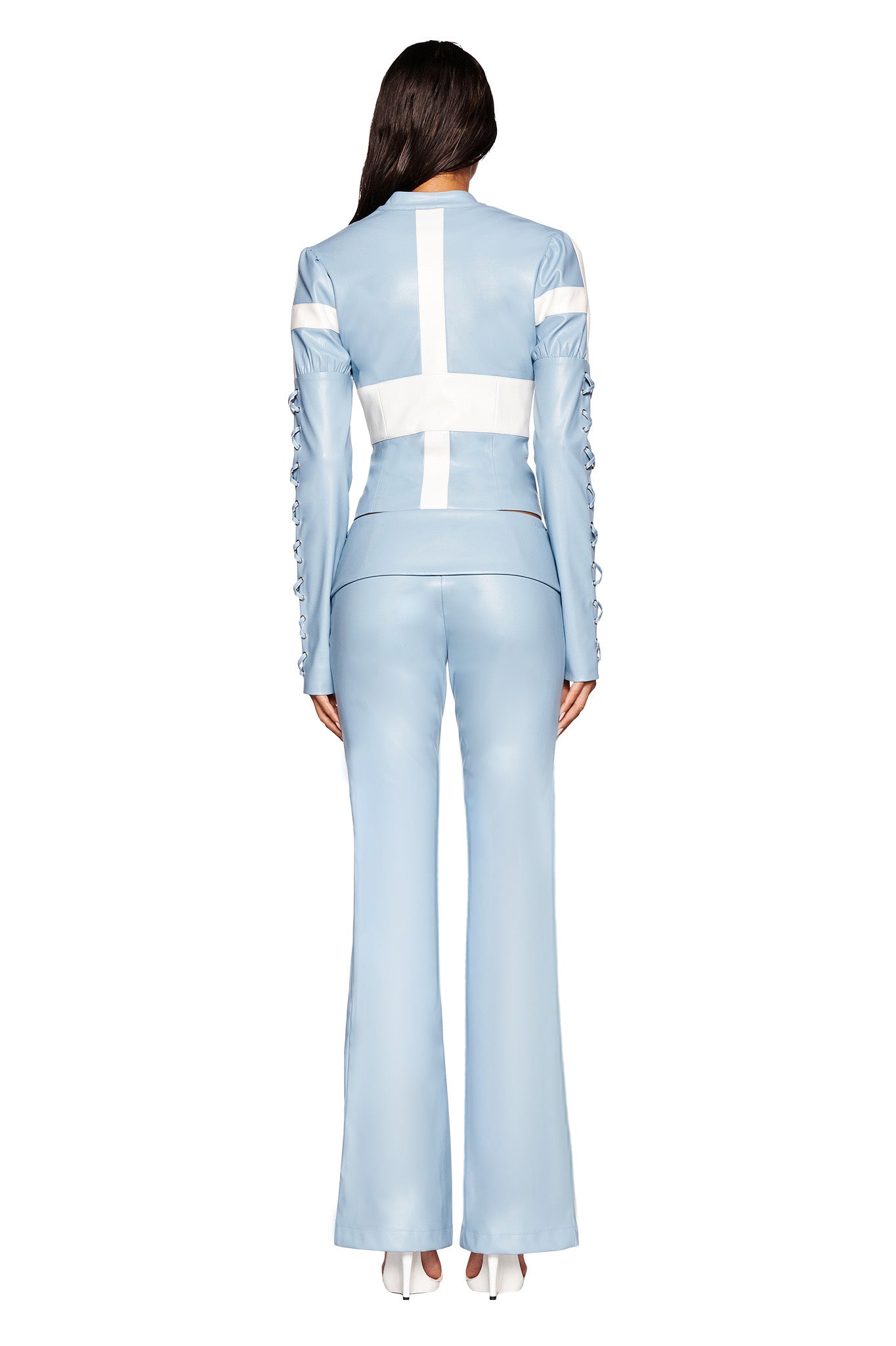 A woman stands facing away, modeling the AMALTHEA PANT - BLUE from I.AM.GIA in baby blue satin with a white cross design on the back. The suit features a fitted blazer adorned with decorative sleeve buttons and high-waisted flared leg pants. She completes the look with white high heels.
