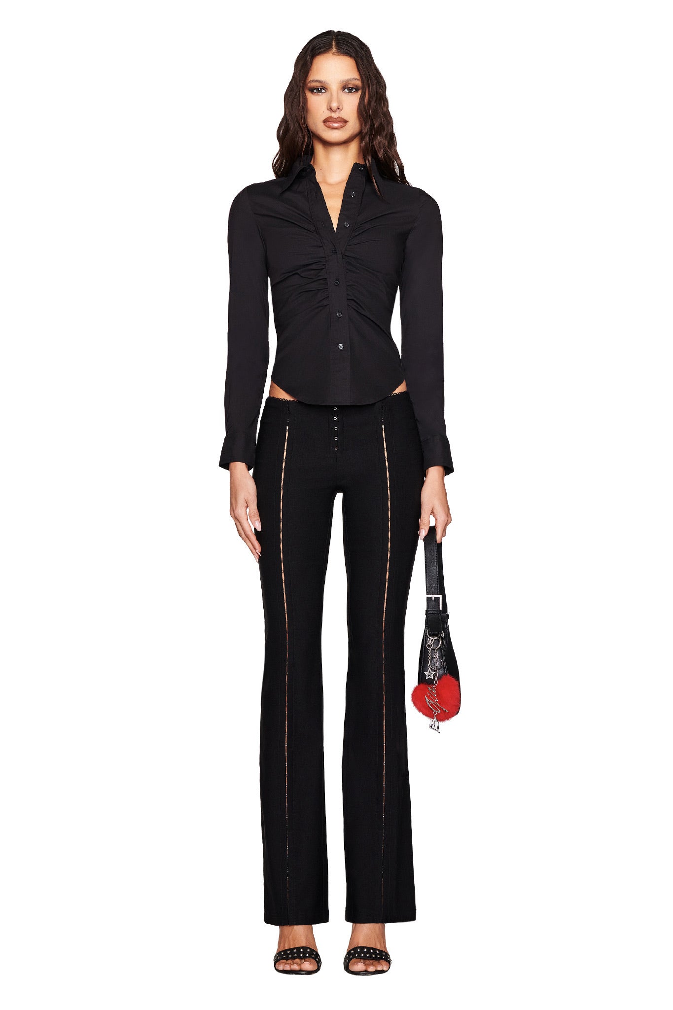 A woman with long wavy hair wears the I.AM.GIA MONICA SHIRT - BLACK, stylish black pants with decorative seams, a black and red purse, and sleek black high-heeled sandals.