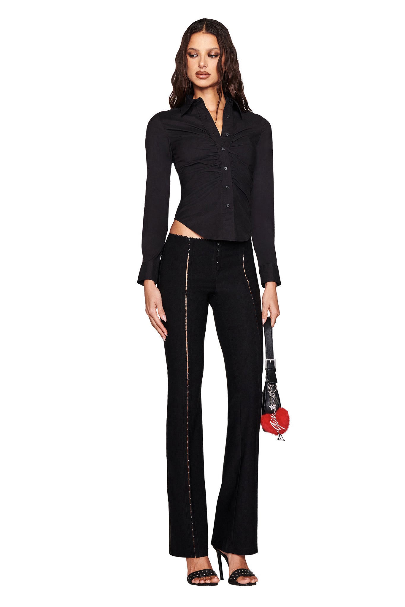 Dressed in the MONICA SHIRT - BLACK by I.AM.GIA and flared black pants with zipper detailing, a person poses holding a handbag with a red heart accessory. Their long, wavy hair contrasts against the plain white background.