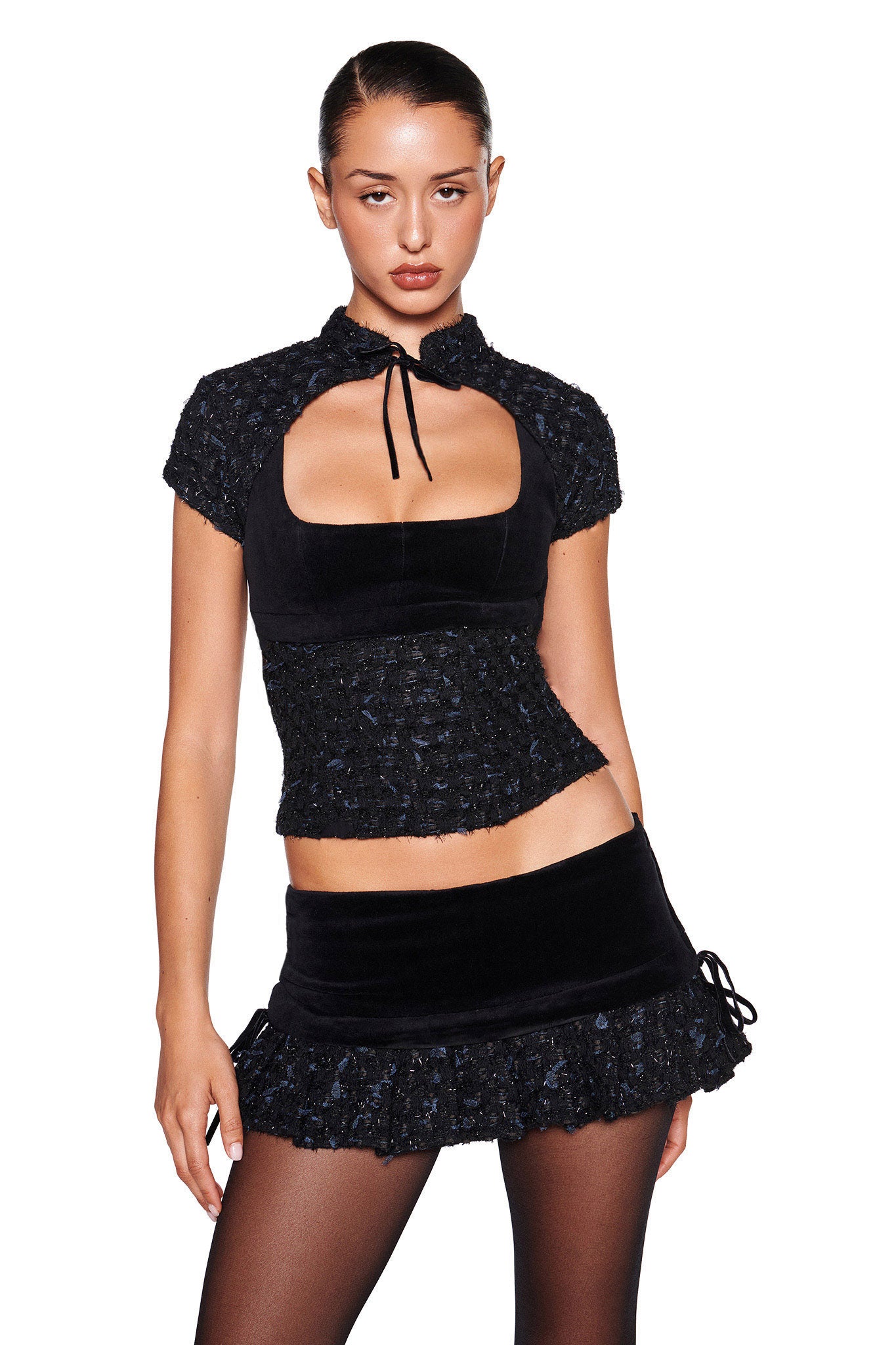 A person stands against a plain white background, dressed in the stylish SAFFRON 3 PIECE CORSET - BLACK by I.AM.GIA. The outfit showcases a short-sleeved top featuring a unique rectangular cutout in the front with a tie at the neckline, complemented by a ruffled mini skirt that mimics the texture of boucle. The person has their hair pulled back.