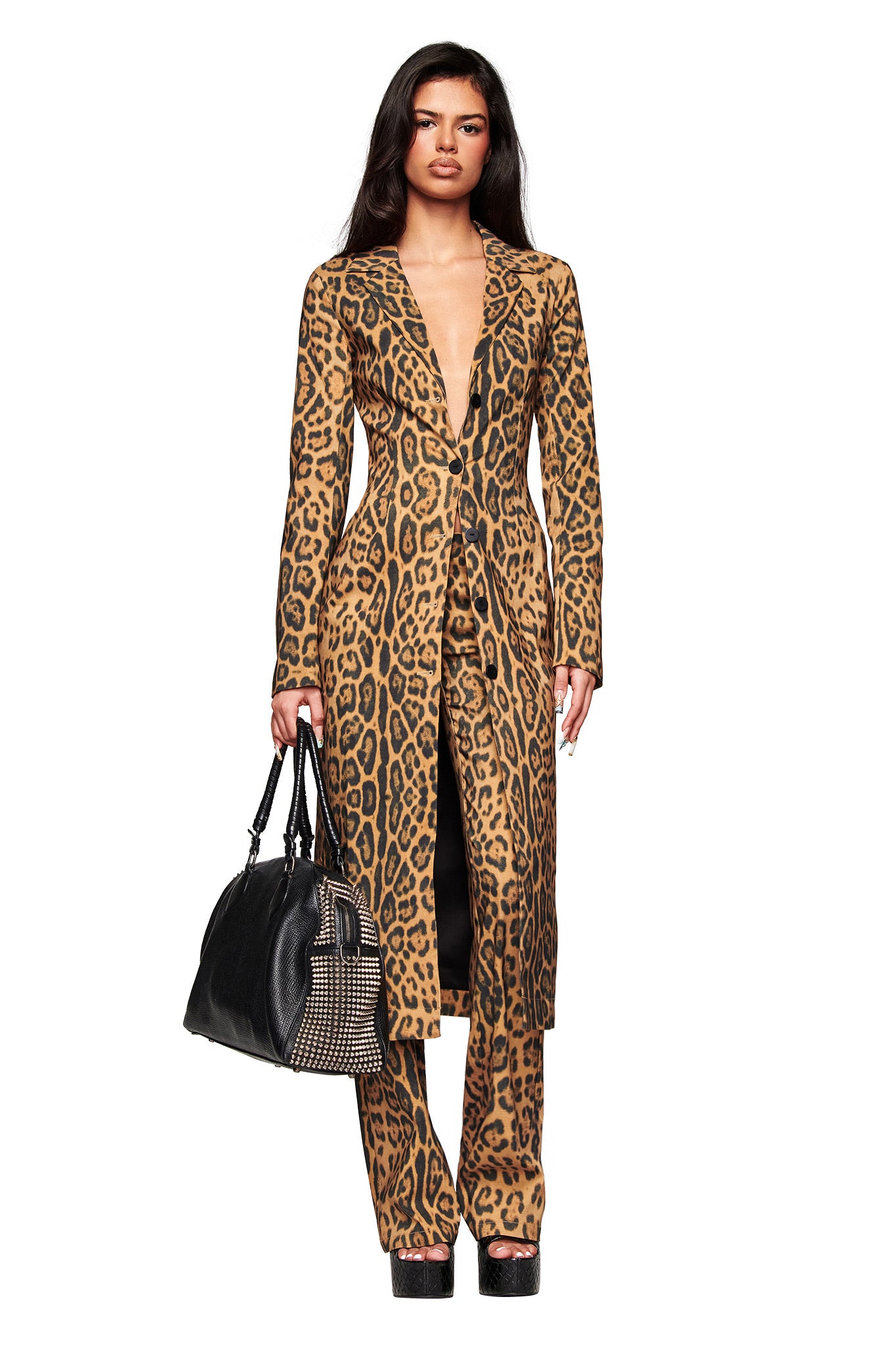 A woman wears the I.AM.GIA UMA COAT - PANTHERA with matching Panthera print pants, carries a large black handbag, and stands on platform shoes against a white background.