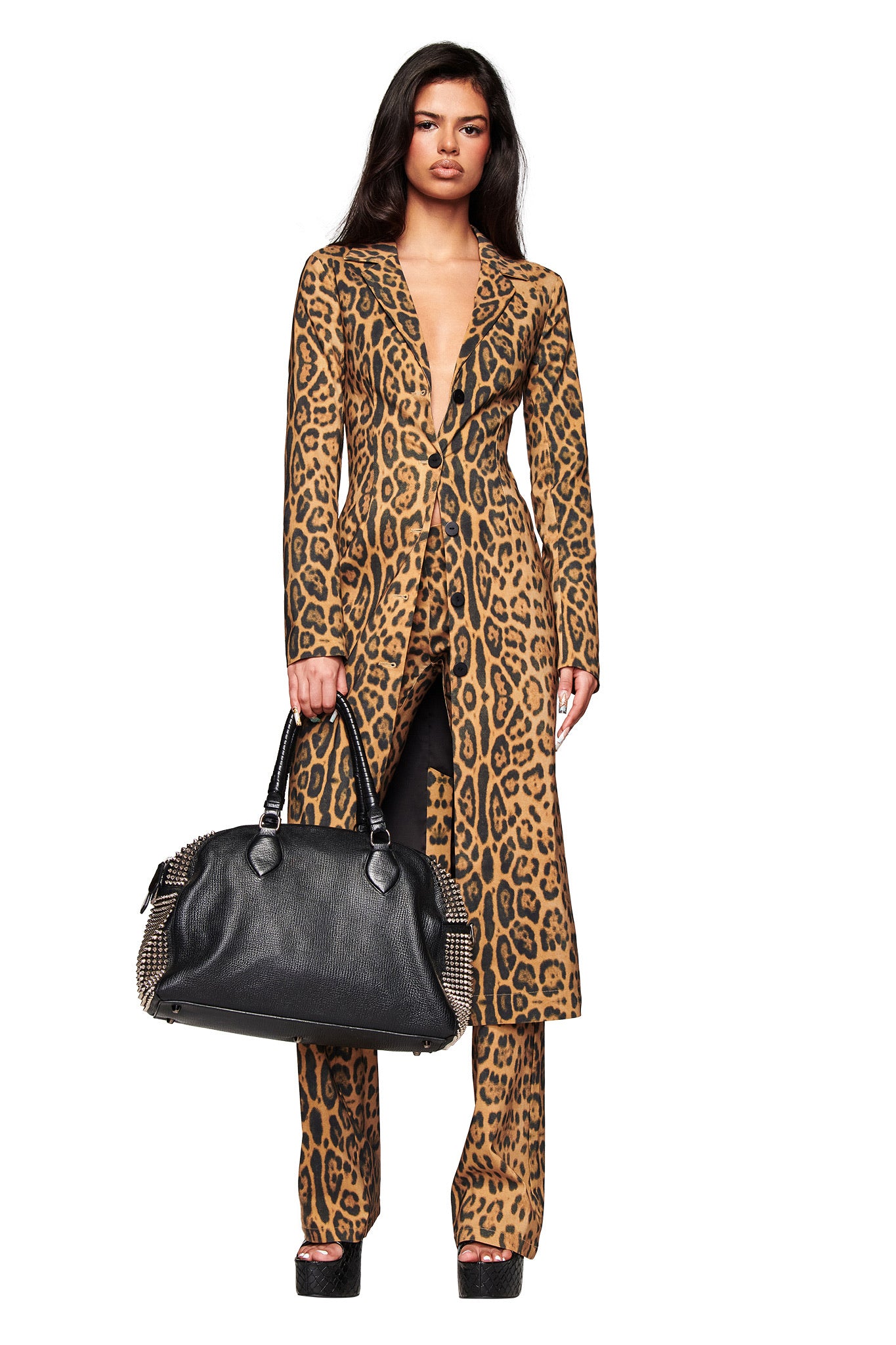 A woman with long dark hair, neutral expression, and platform heels faces forward against a white background, wearing the I.AM.GIA UMA COAT - PANTHERA and matching pants in animal print, and holding a large black handbag.
