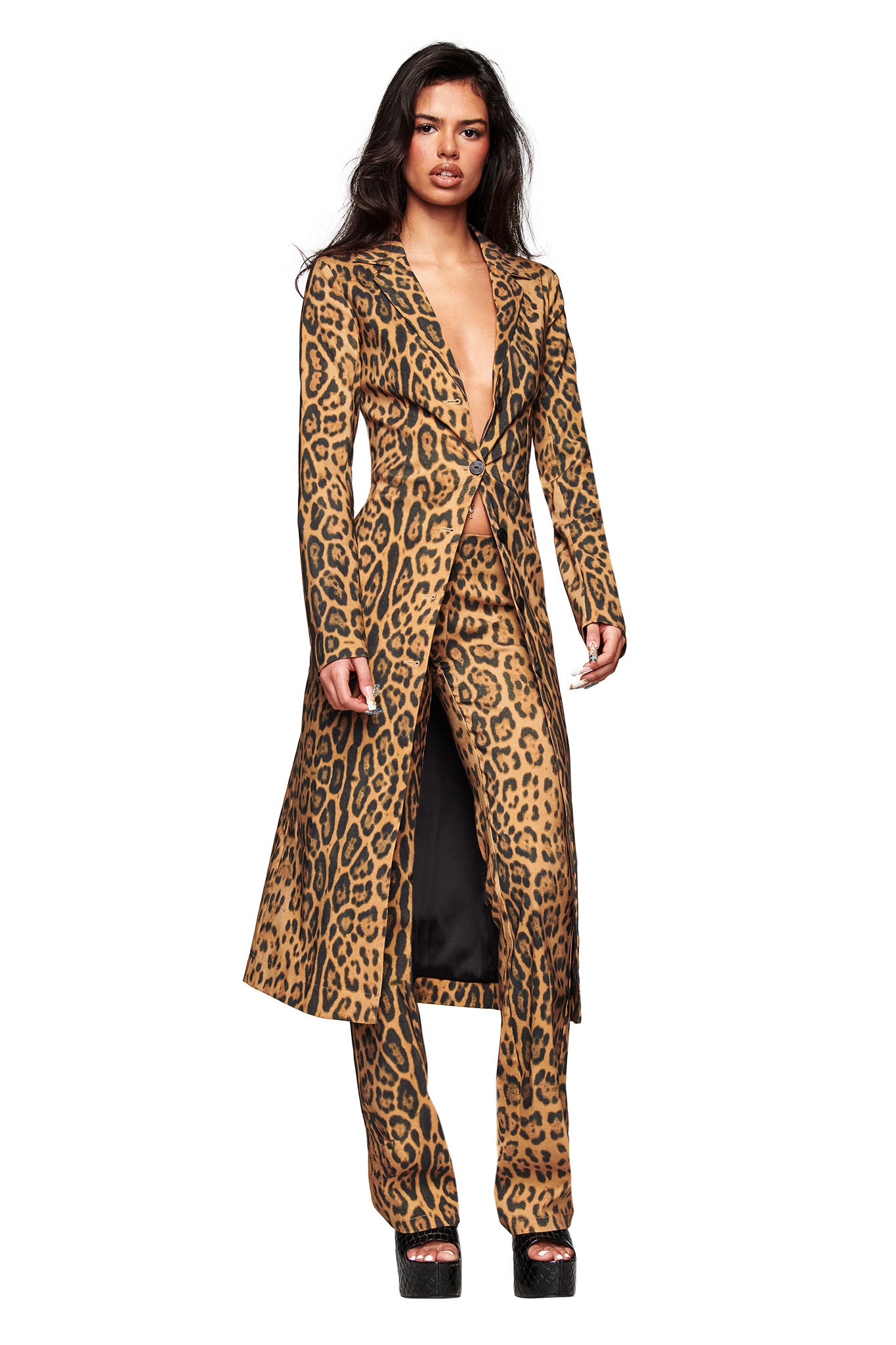 A woman poses confidently against a white background in the I.AM.GIA UMA COAT - PANTHERA, a tailored longline animal print coat styled open at the chest with matching pants and platform heels.