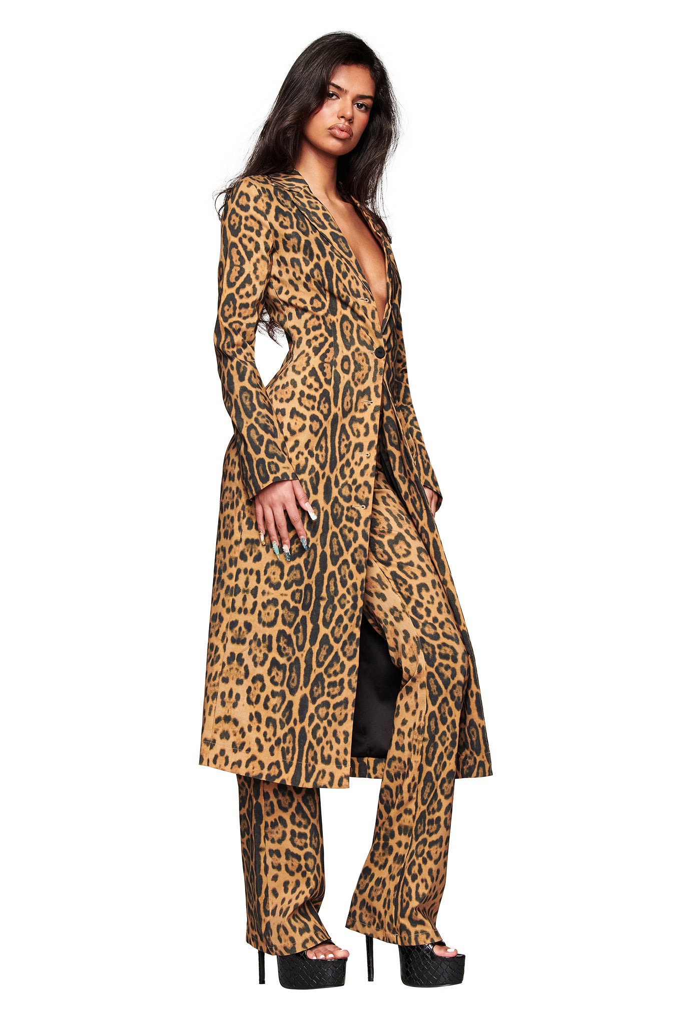 A woman models the I.AM.GIA UMA COAT - PANTHERA, a tailored animal print longline coat, paired with matching pants and black platform heels. She stands against a white background, gazing confidently at the camera.