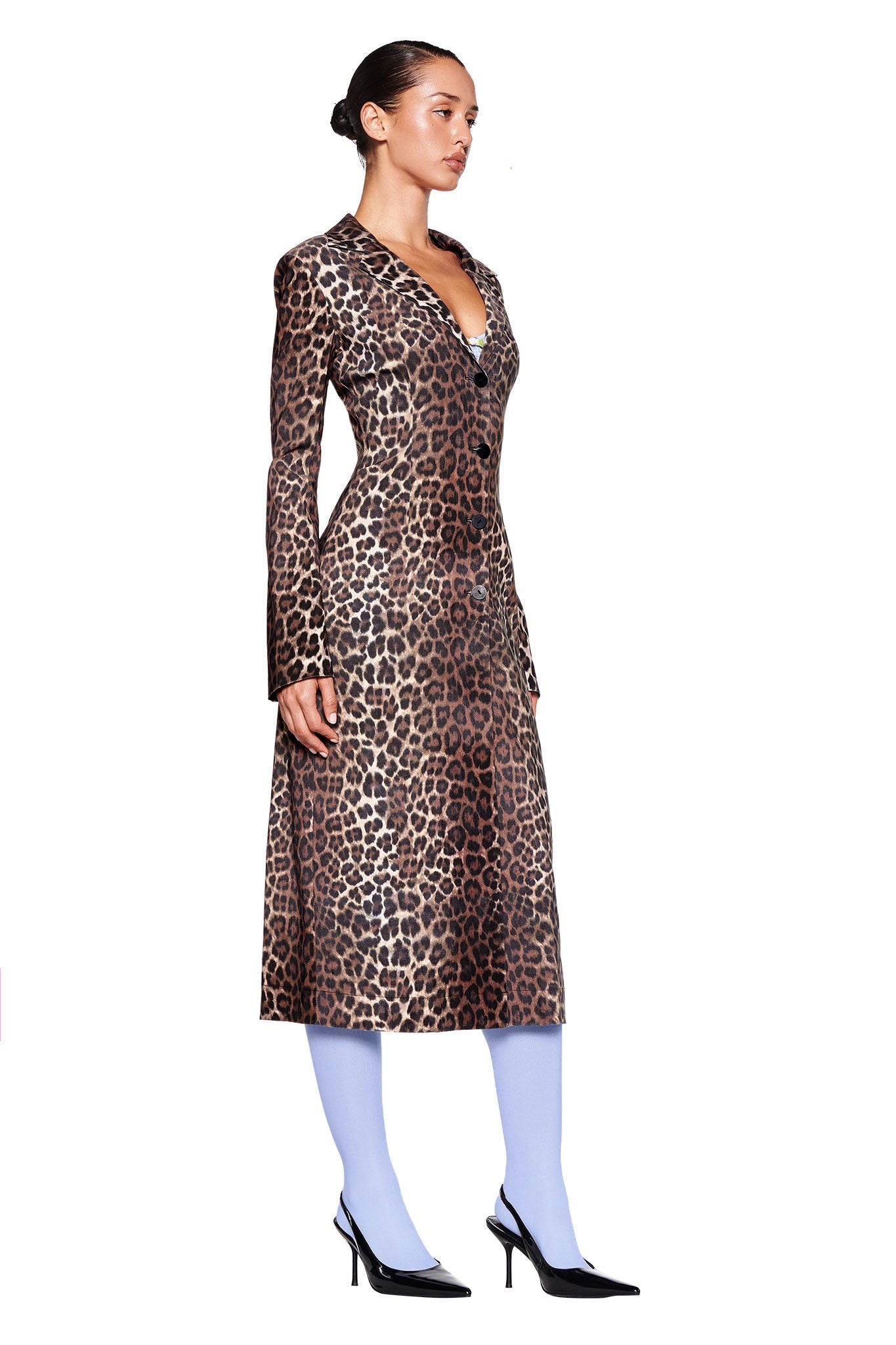 A woman stands in profile against a white background, wearing the long, tailored UMA COAT - NEUTRAL : LEOPARD by I.AM.GIA. She pairs it with light blue tights and black high-heeled shoes, her hair pulled back in a neat bun.