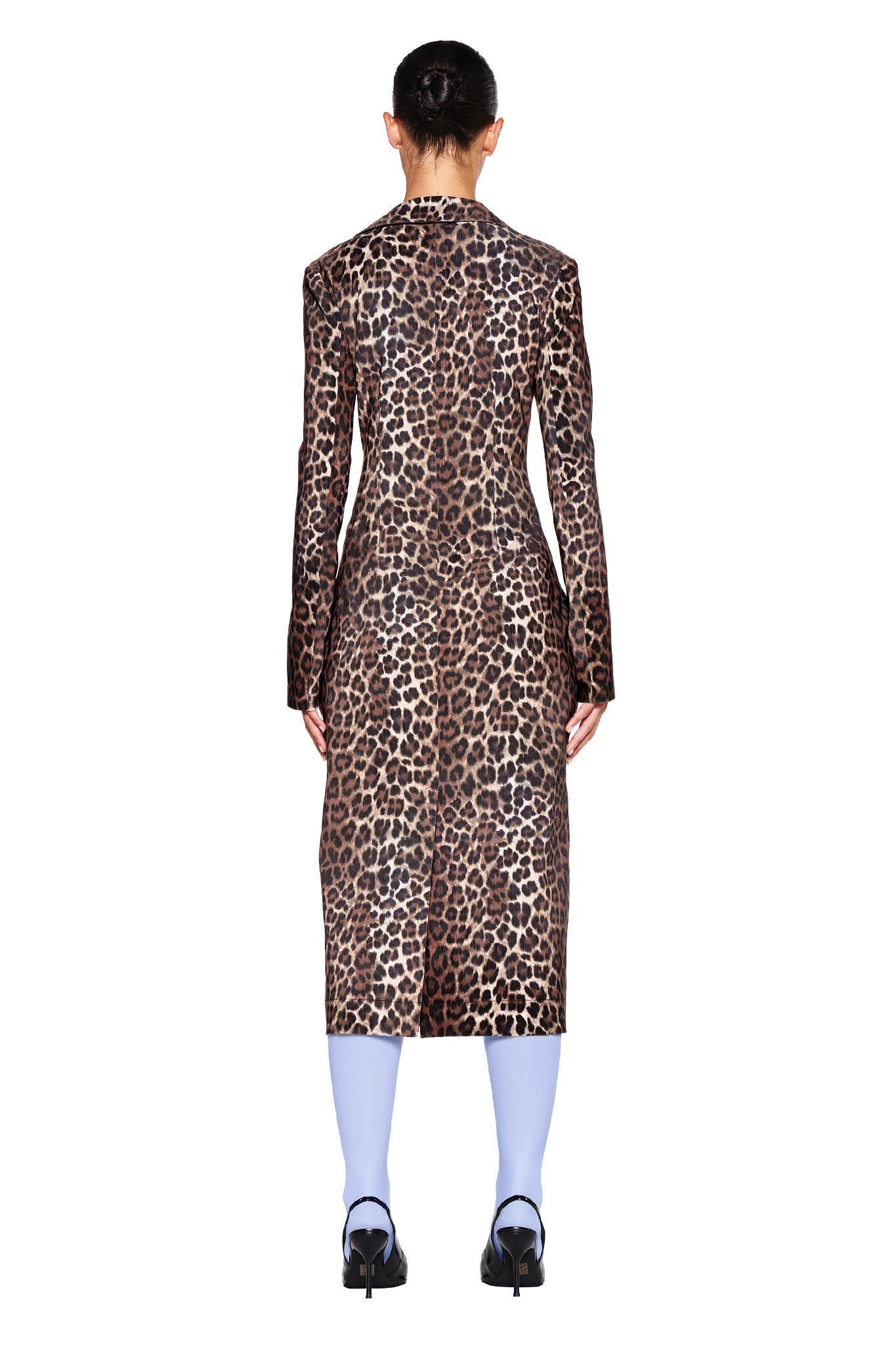 A person with their back to the camera is wearing the UMA COAT - NEUTRAL : LEOPARD by I.AM.GIA, a tailored leopard-print coat that extends below the knees in a stylish midi length. They are also wearing light blue socks and black high-heeled shoes. Their hair is styled in a bun, set against a white background.